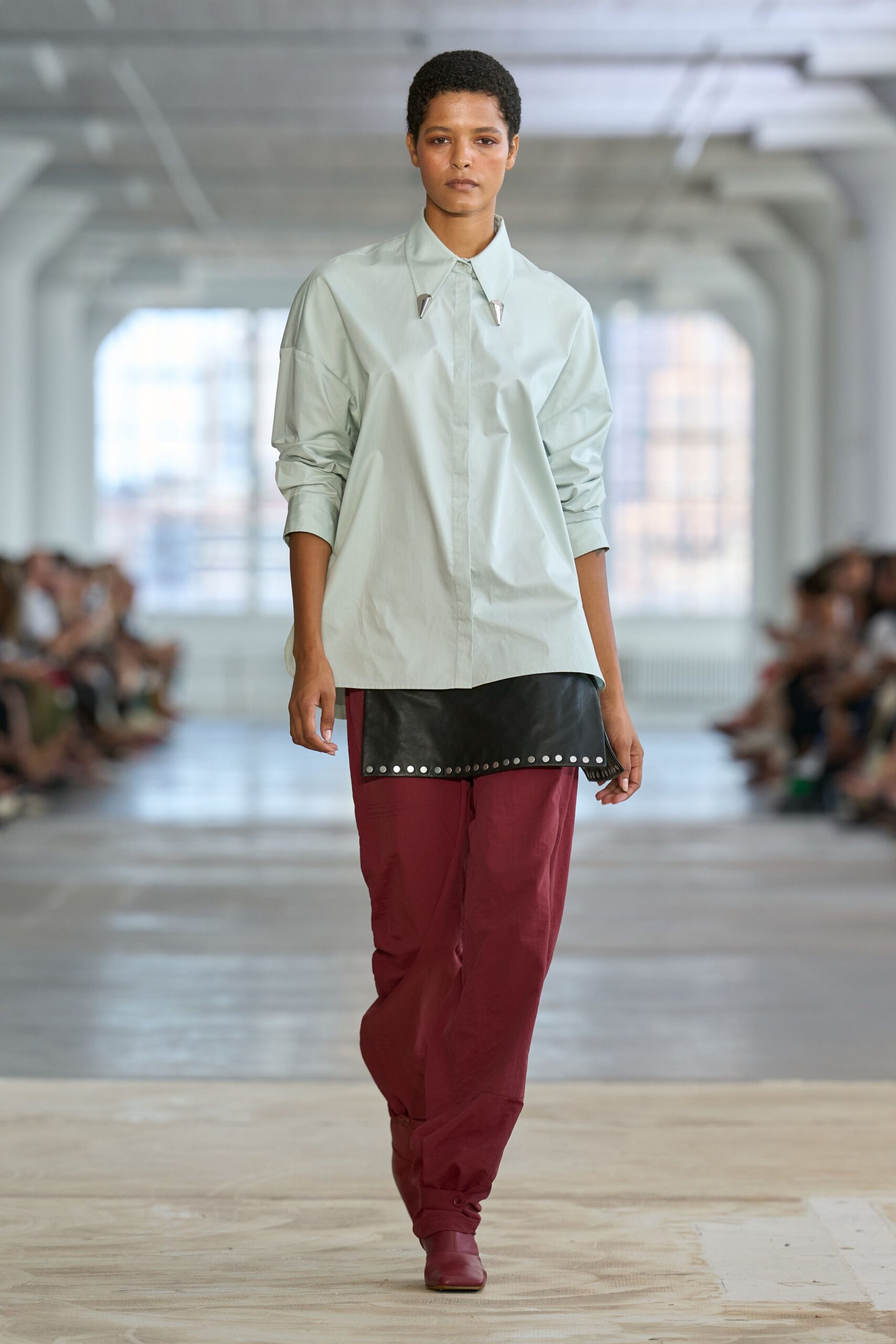 Pale celadon cotton shirt, dropped shoulders and two silver collar tips. Black leather apron panel, hem punched with neat silver studs. Oxblood trousers gathered at the ankle with a tiny button tab. Square-toe oxblood leather boots. For crossing a museum atrium before the rain starts again.