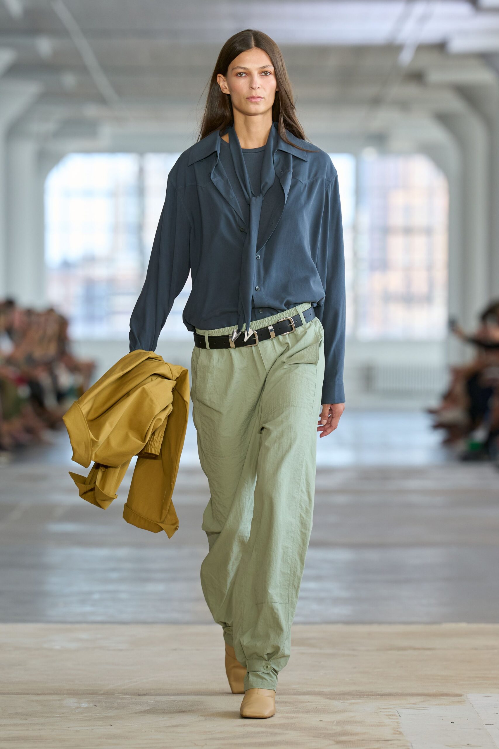 Slate silk blouse with a long scarf tie knotted loose. Black leather belt with oversized silver tip and keeper. Pale sage nylon trousers, elastic waist and buttoned ankle tabs. Square-toe beige leather boots. Mustard cotton jacket carried folded over one hand. For crossing town after rain with dry cleaning on your arm.