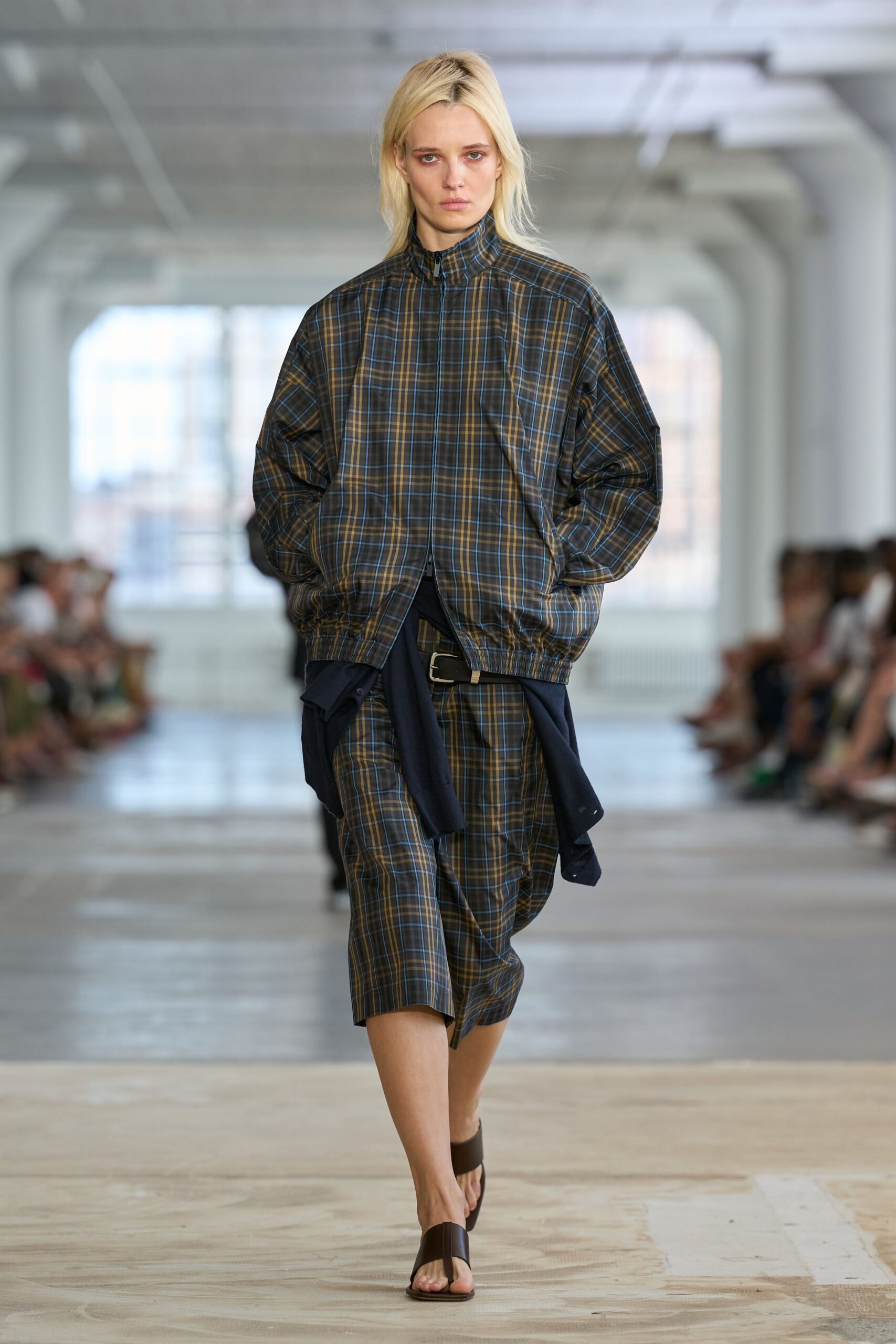 Brown plaid track jacket in crisp nylon, zip stopped at the throat. Navy button-front shirt tied loose around the hips. Black leather belt with a square silver buckle. Matching plaid knee shorts, cut roomy through the leg. Dark brown thong sandals. Office park courtyard, lunch break gone long in August heat.