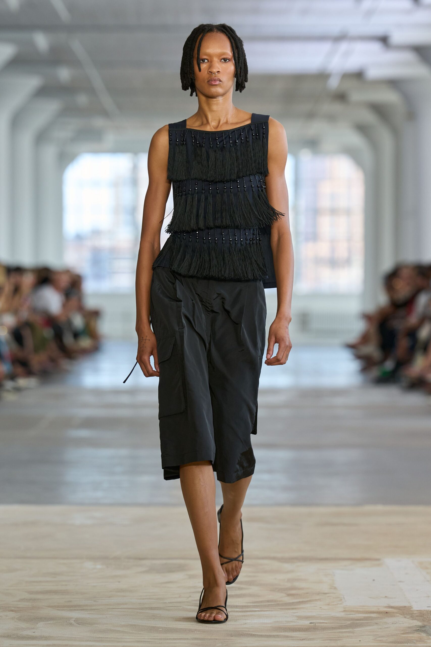 Black sleeveless top banded with three tiers of beaded fringe. Matte black wrap skirt in crisp technical cloth, one cord end dangling. Bare black lace-up sandals. Crossing a museum atrium after hours, hearing your steps answer back.