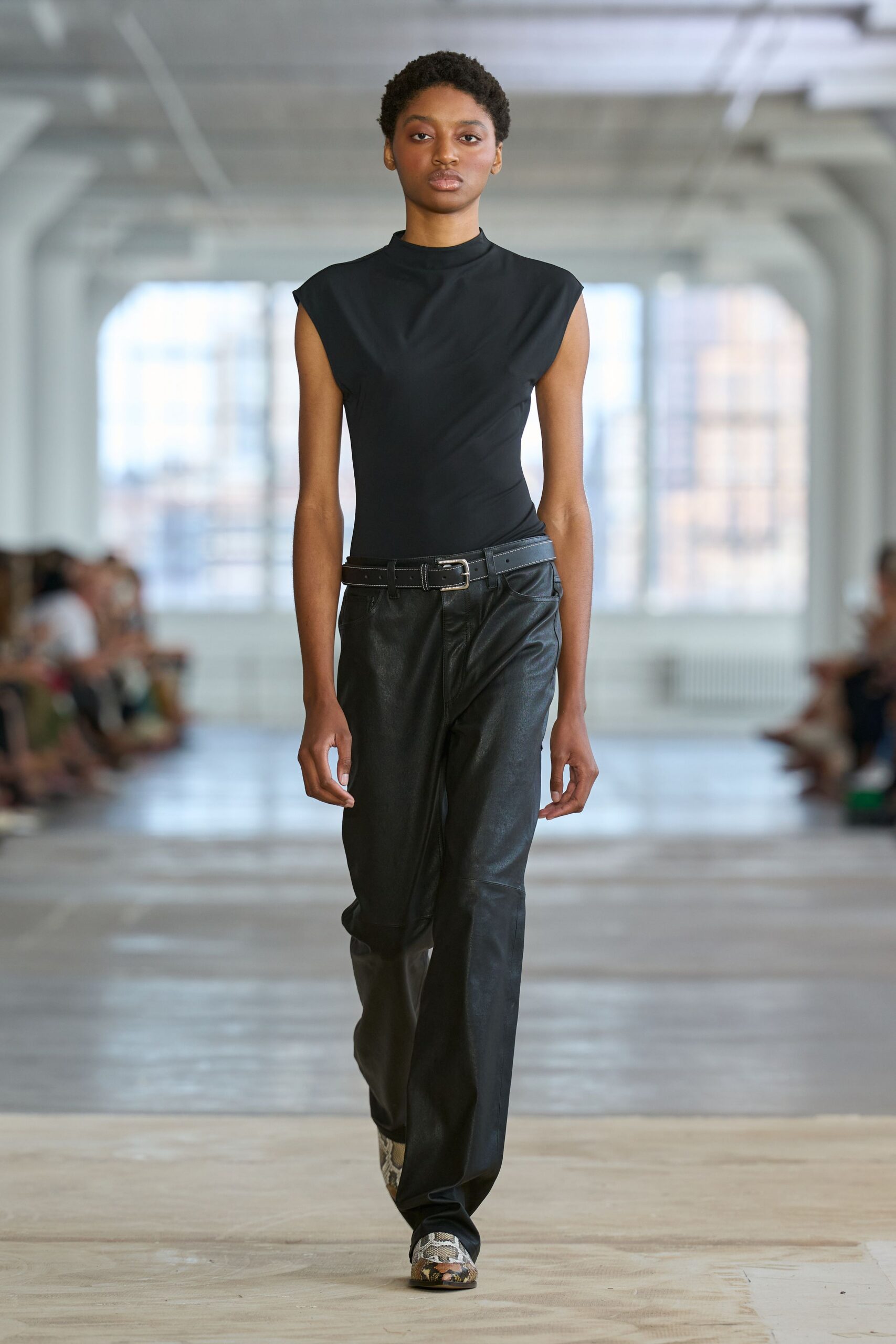 Black stretch jersey shell, cut with a close mock neck and cap sleeves. Black leather belt with a square silver buckle sitting slightly askew. Black leather trousers, loose through the leg with a soft break. Python boots peeking out under the hem. Office tower Sunday, nobody else on the elevator but the cleaner.