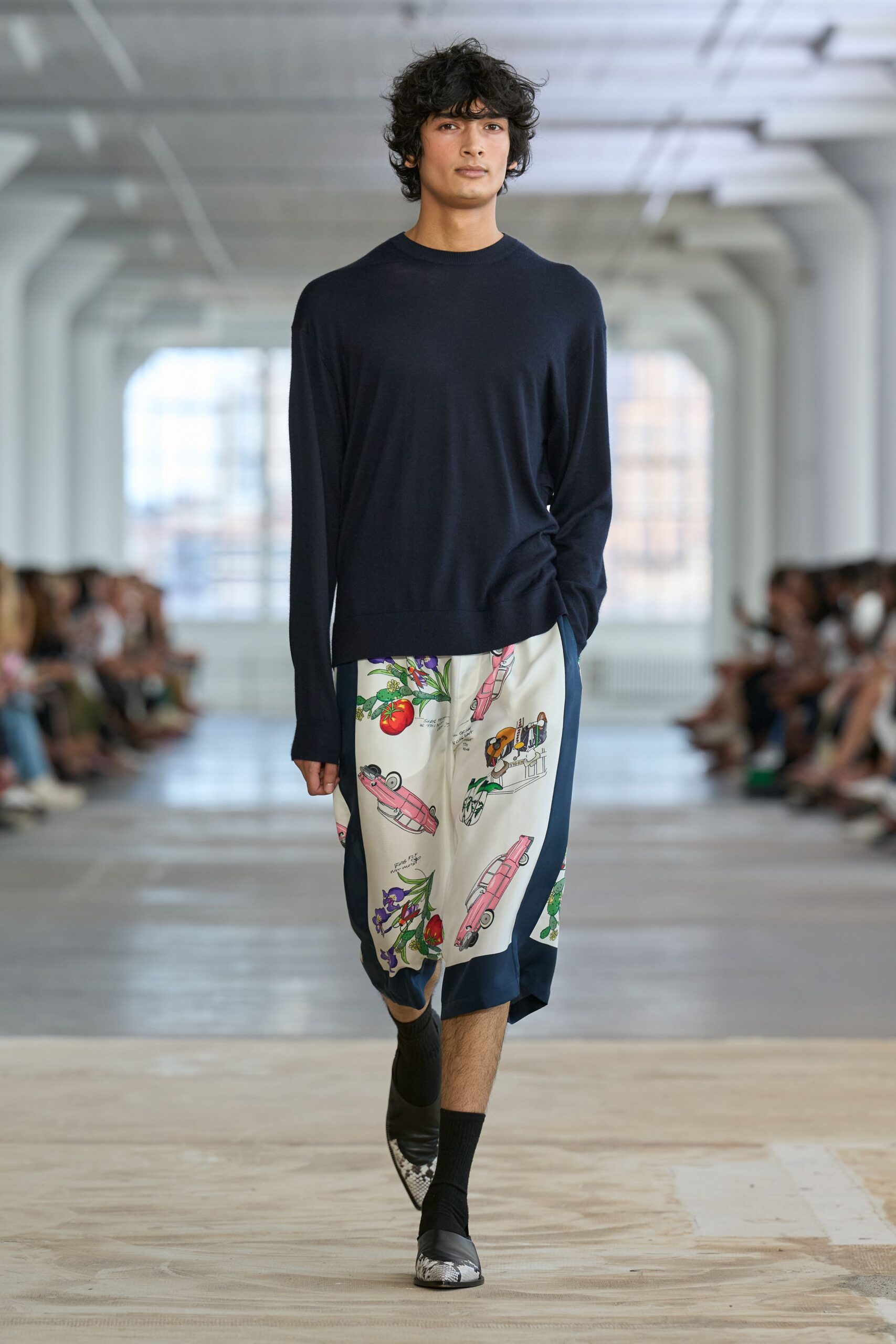 Ink navy fine-gauge sweater with a narrow ribbed crewneck. Cream silk panel shorts printed with pink cars and botanical sketches, navy sides dipping longer. Black ribbed socks. Pointed loafers tipped in grey python. Sunday museum cafe, sketchbook open beside a melting espresso.