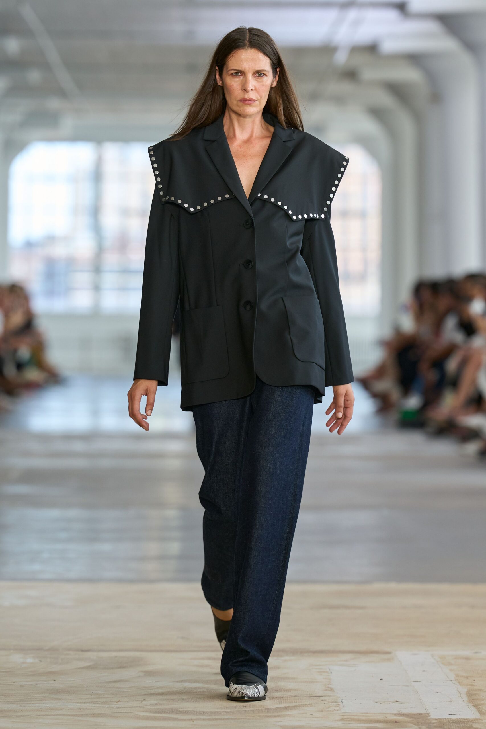 Black blazer with an oversized sailor collar ringed in silver studs. Dark indigo denim trousers cut loose through the leg. Black slip-ons with scuffed white rubber at the toe. Crossing an empty ferry terminal before the first announcement crackles on.