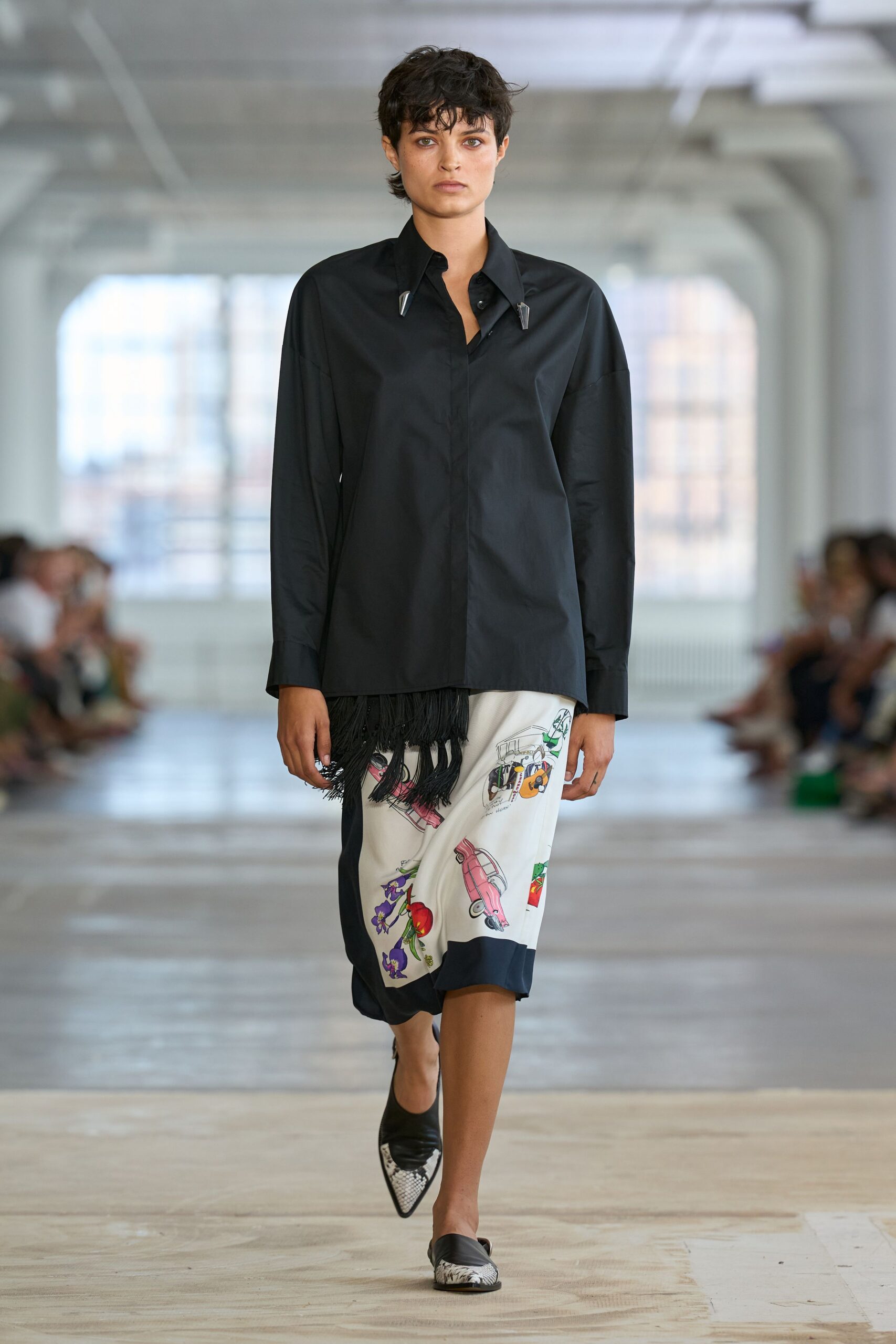Black cotton poplin shirt with chrome collar weights and a fringe hem. Cream satin midi skirt printed with pink cars and scattered produce. Pointed black leather mules tipped in python-print panels. Walking past closed fruit stalls with yesterday's market flyers underfoot.