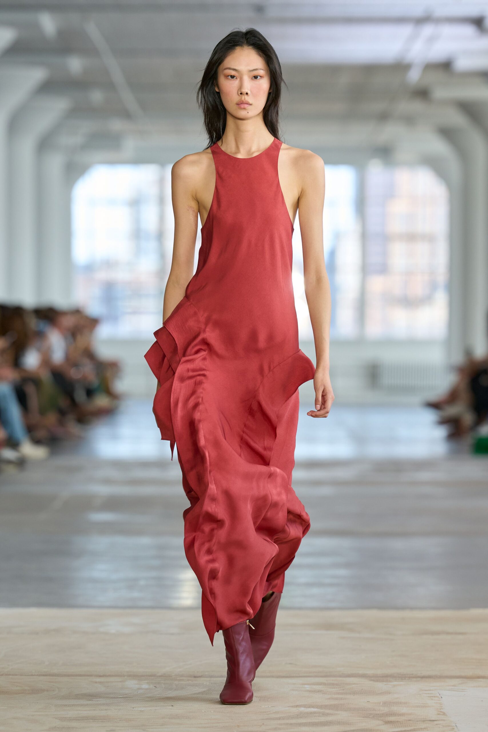 Brick-red satin dress with a high racer front and sculpted hip flounces. Matching oxblood leather boots, narrow at the ankle. Crossing an empty hotel lobby before sunrise, flowers from last night still breathing.