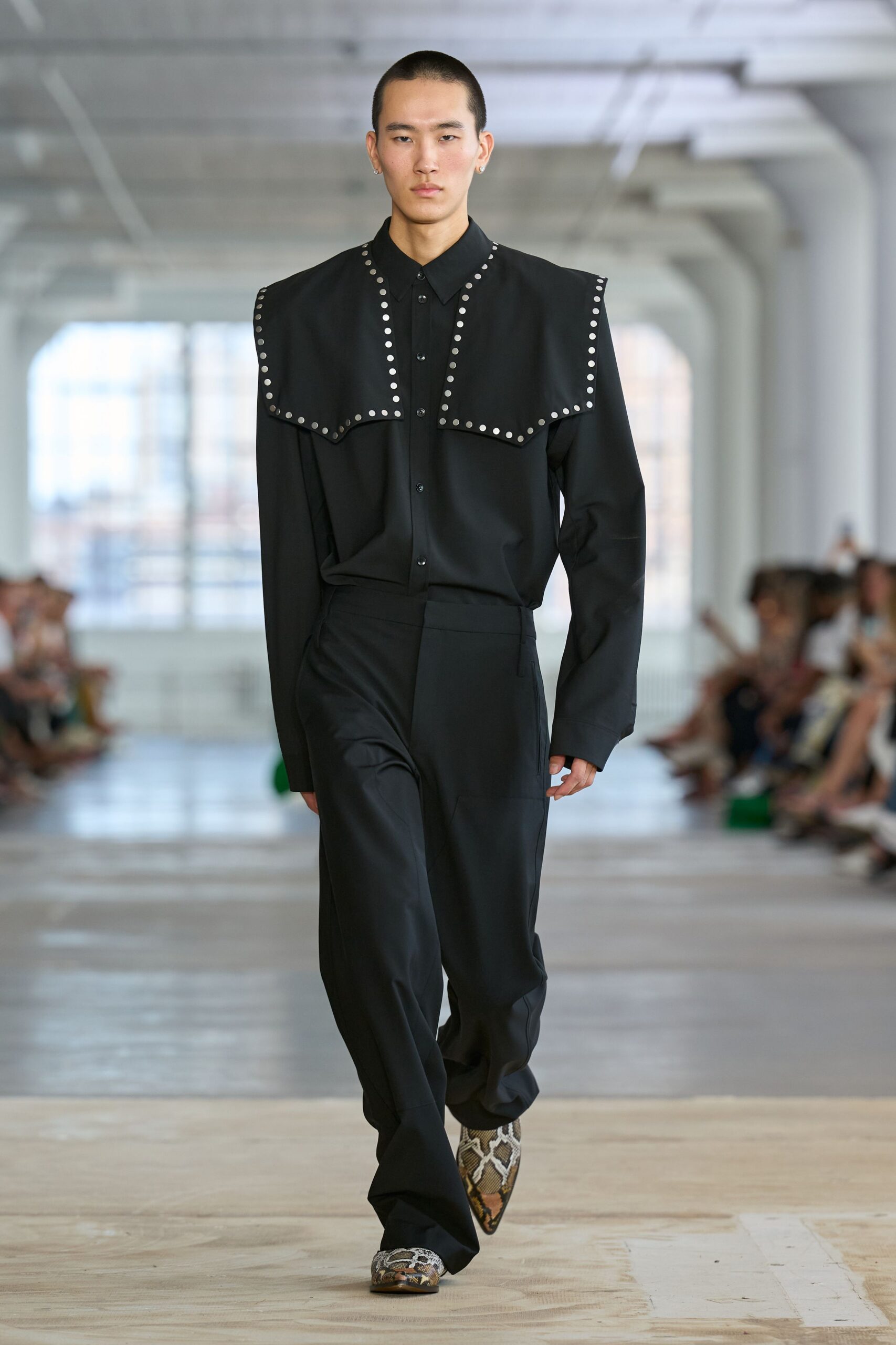 Black button-front shirt with a broad studded cape collar angling over the chest. High-waisted black trousers falling long enough to pool over the foot. Pointed python boots with a pale vamp and darker mottled toe. Walking the hotel corridor carpet after midnight ice has melted from the cuffs.