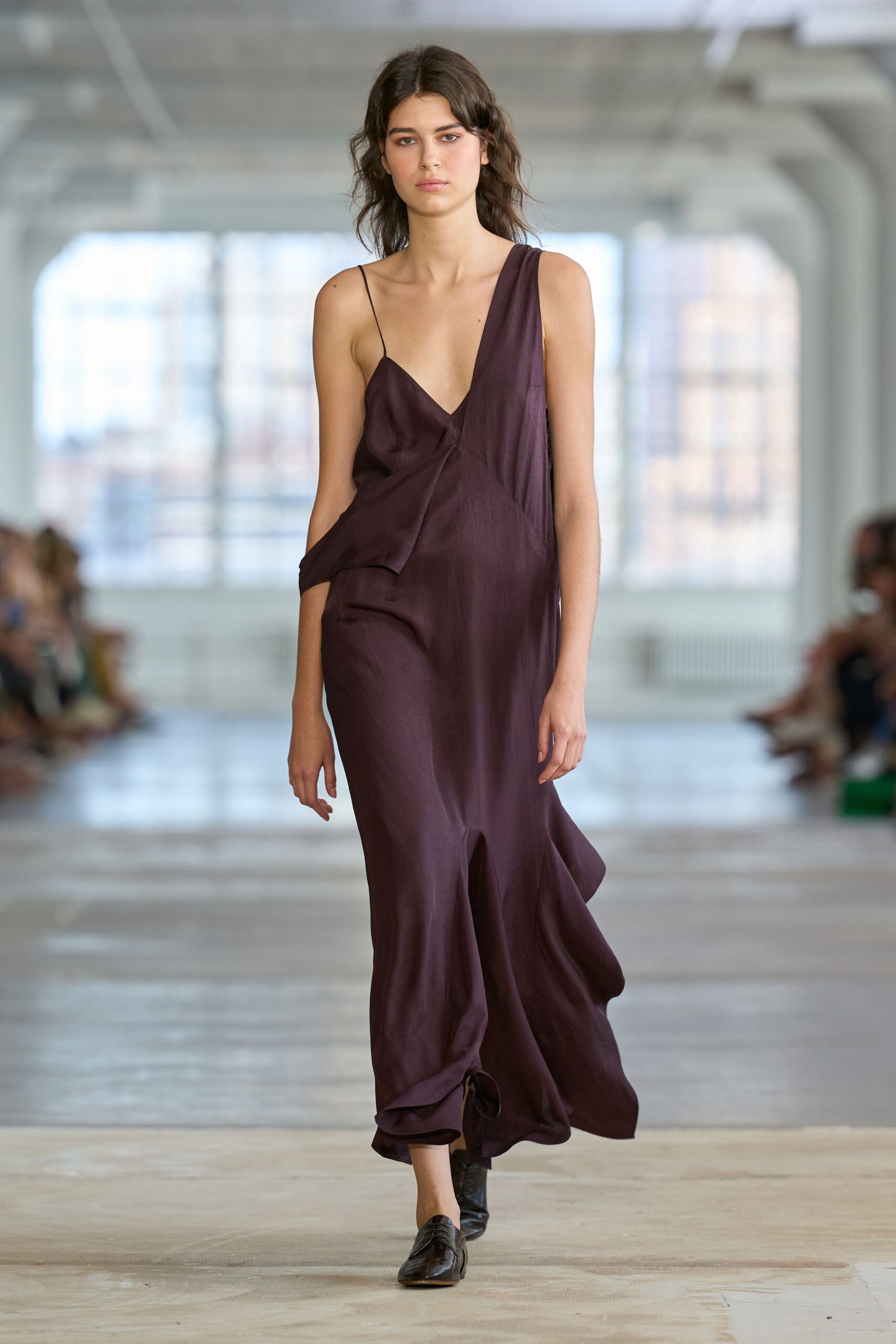 Aubergine satin slip dress with one thin strap and a folded side drape. Black square-toe flats, softly creased across the vamp. Morning after the wedding, crossing the hotel lobby for coffee.