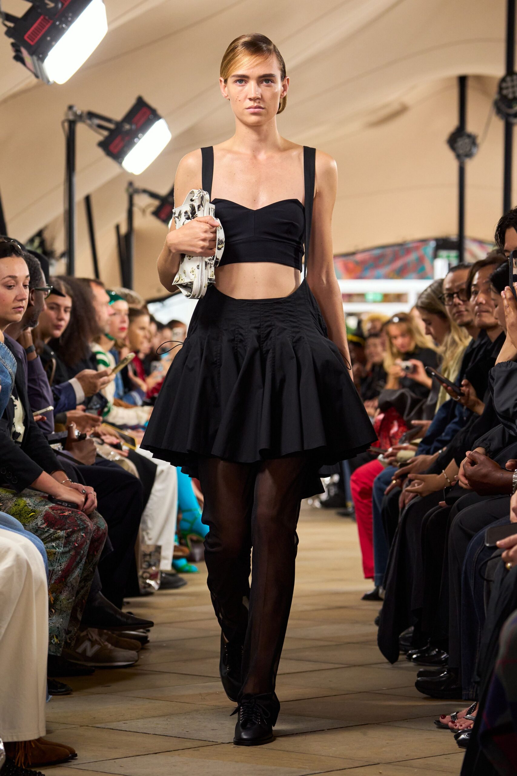 Black structured crop top with a sweetheart curve and wide straps. Black drop-waist skirt cut into a puffed mini overskirt. Sheer black tights. Black lace-up oxfords. White floral clutch tucked hard under one arm. Crossing a museum courtyard before the fountains switch on.