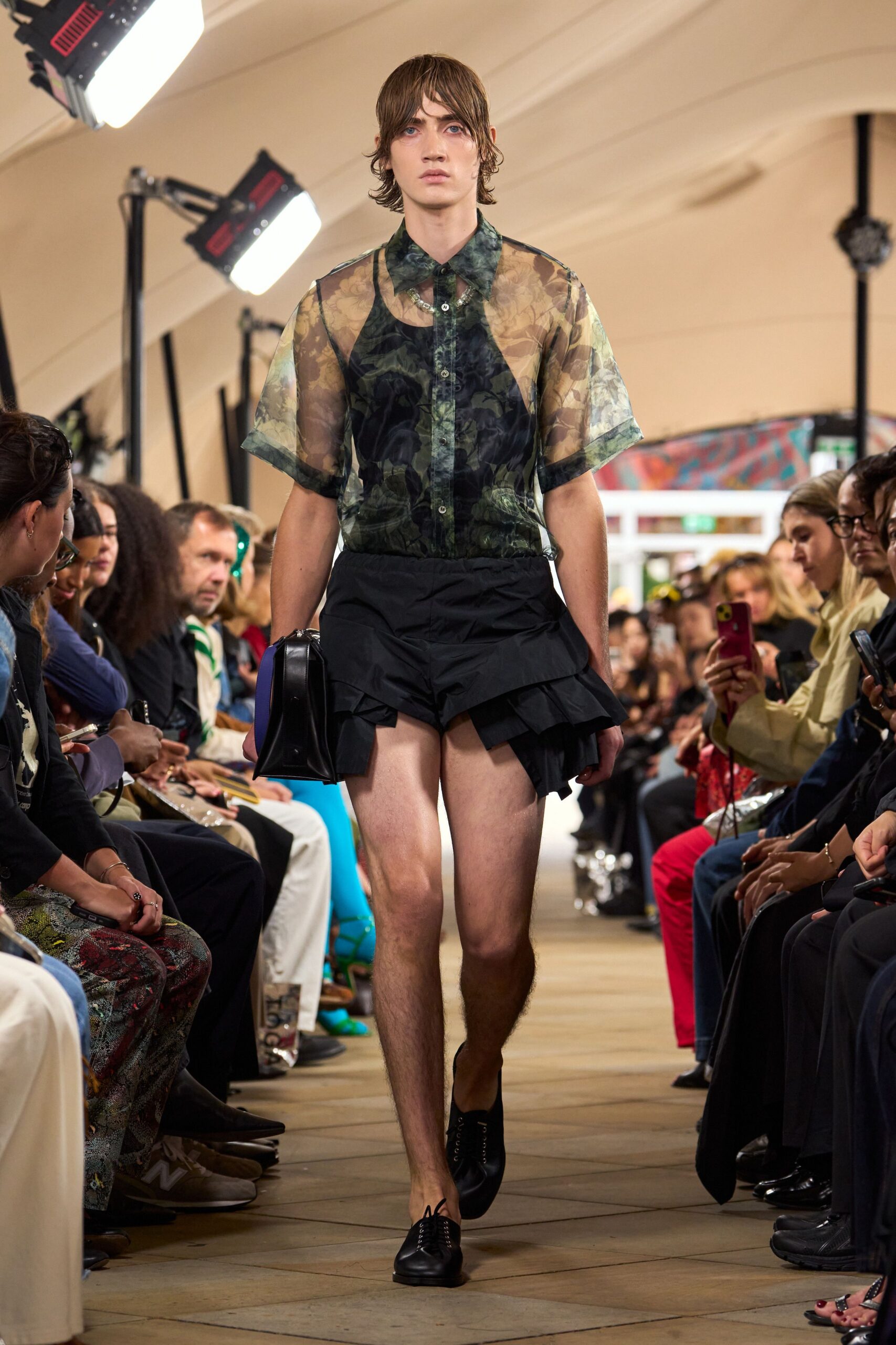 Transparent floral organza shirt, dark tank shape visible beneath the placket. Black shorts with a wrapped peplum ruffle kicked up at one hip. Boxy black leather bag with a cobalt side panel. Black leather lace-ups, narrow and plain. Walking into a midsummer reception before the lawn has dried.