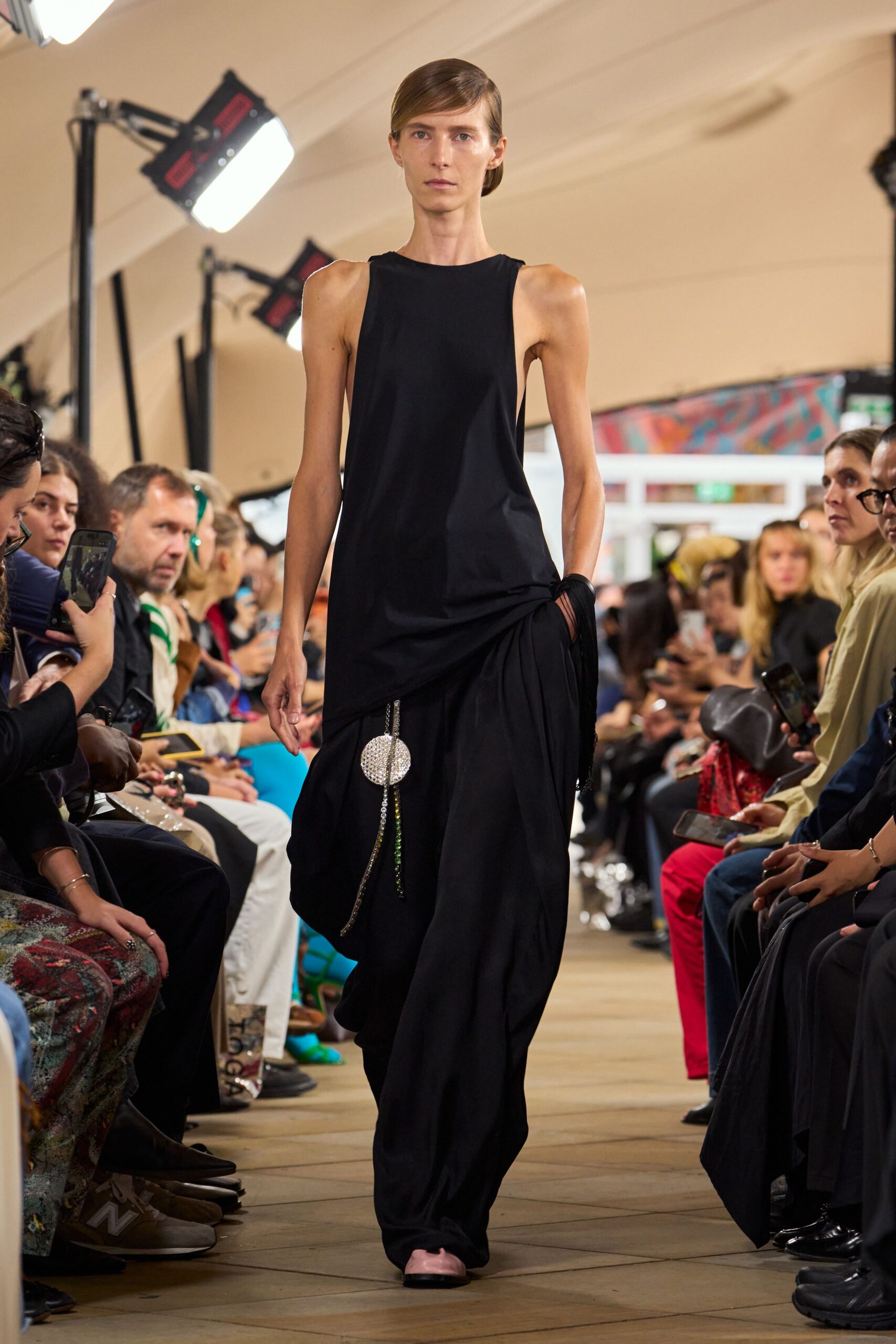 Black jersey column dress with deep armholes and a gathered hip drape. Black fringe-wrapped cuff at the wrist. Crystal disc tassel hanging low from the side seam. Pale pink square-toe flats peeking from the pooled hem. Walking into a museum atrium after the string quartet has packed up.