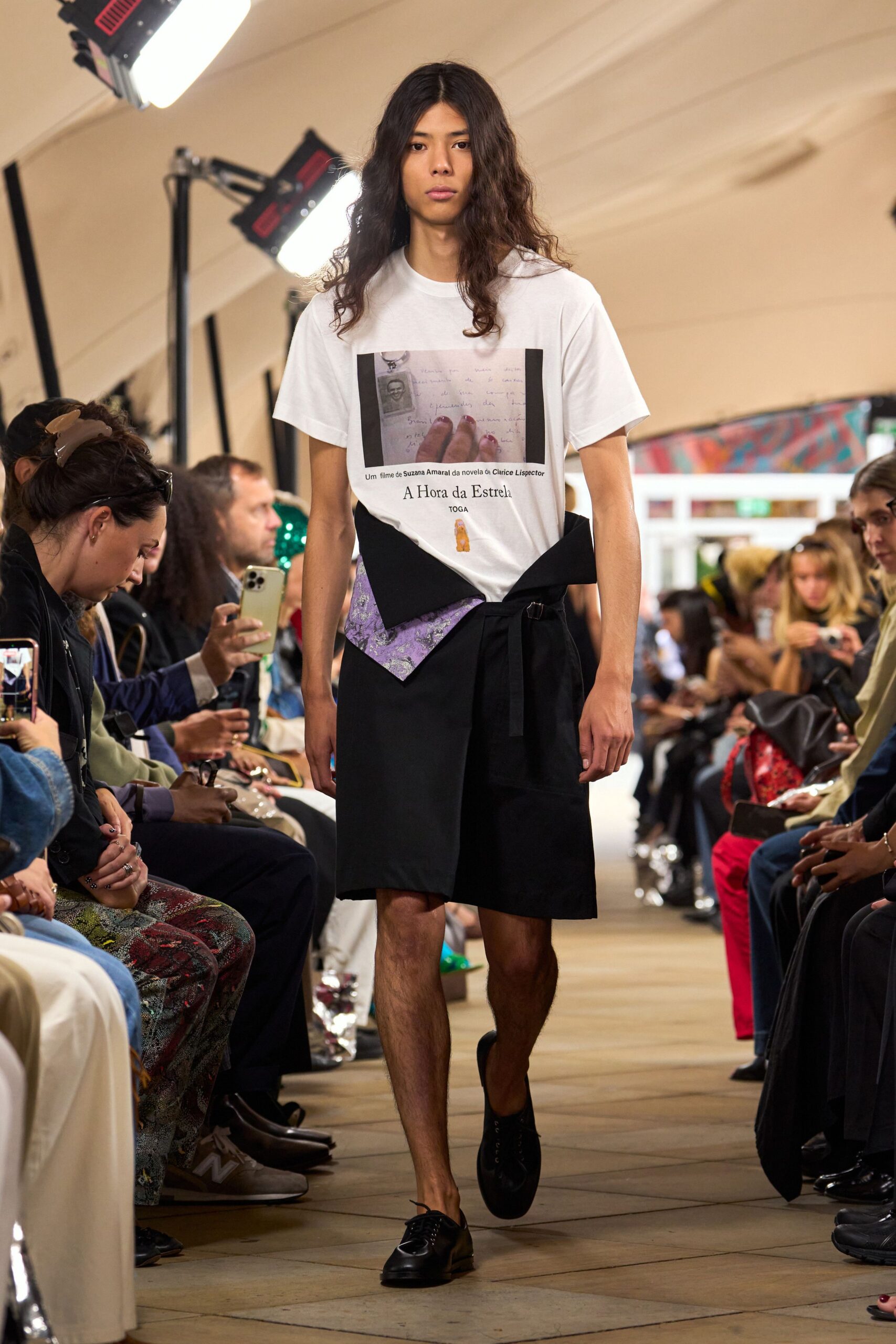 White jersey T-shirt with a film-still print and small serif text. Black wrap shorts tied at the hip, lilac brocade panel peeking out. Black leather derbies with a rounded toe and plain lacing. Walking into a literary festival after missing the first panel.