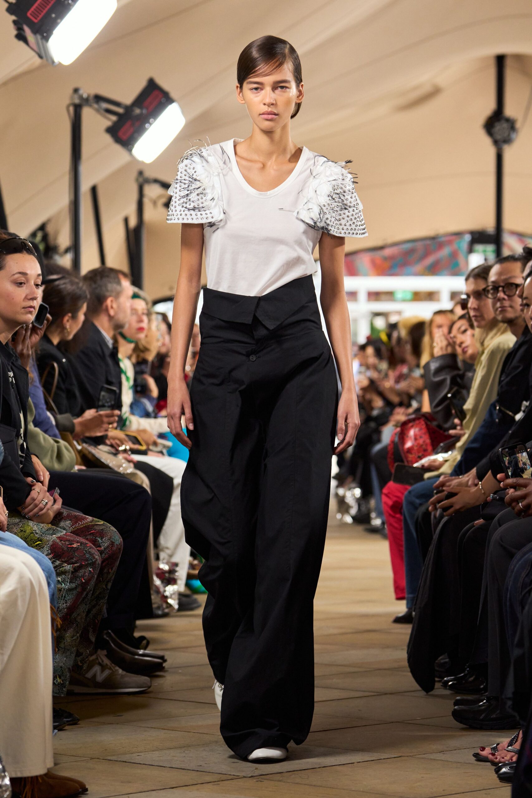 White jersey tee with puffed sleeves crusted in silver studs and wispy feathers. Black wide-leg trousers with an overlapping waistband and one exposed button. White pointed shoes peeking from the pooled hem. Walking into a civic hall luncheon after cutting through rain.