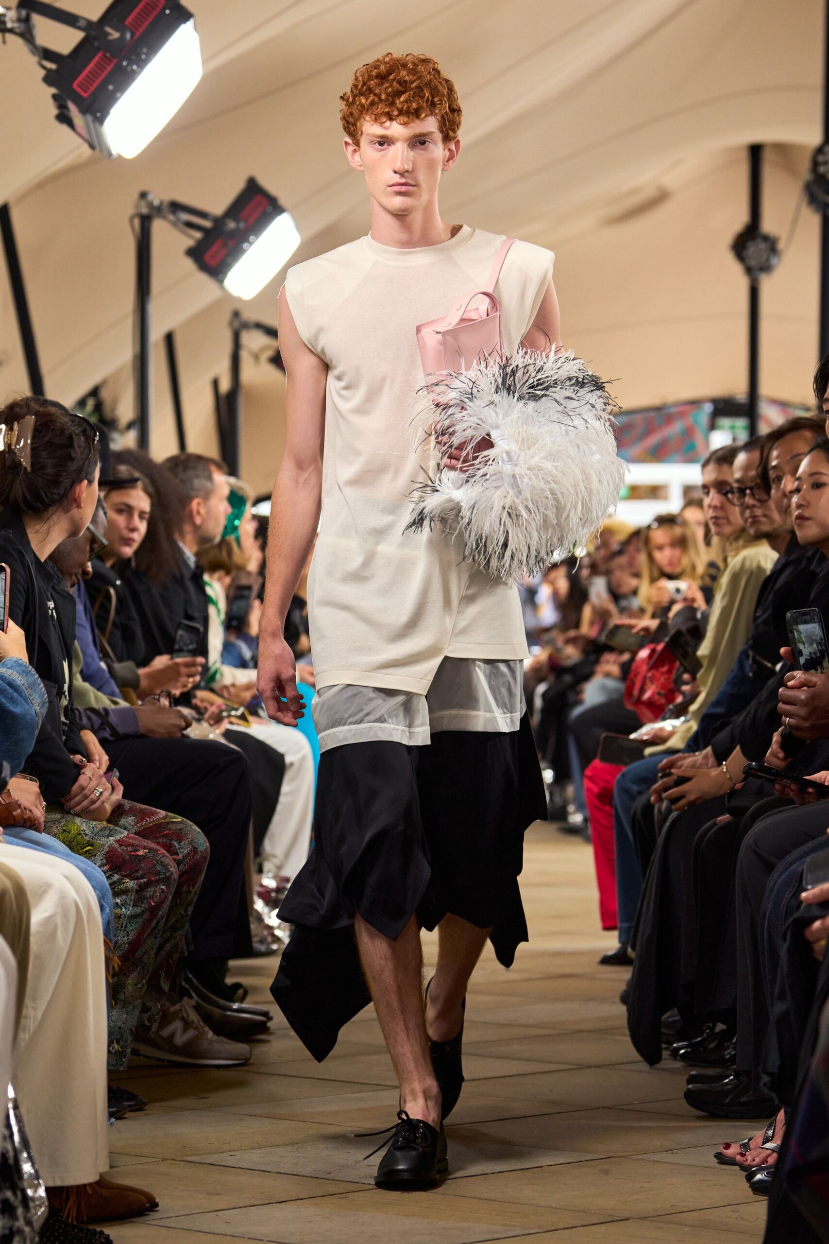 Ivory sleeveless jersey top with a pale pink patch pocket. Glossy blush shoulder bag tucked under a cloud of black-tipped feathers. Black draped skirt layered over a sheer white organza band. Black leather derbies, one lace end trailing loose. Walking into a hotel atrium still carrying the centerpiece from lunch.