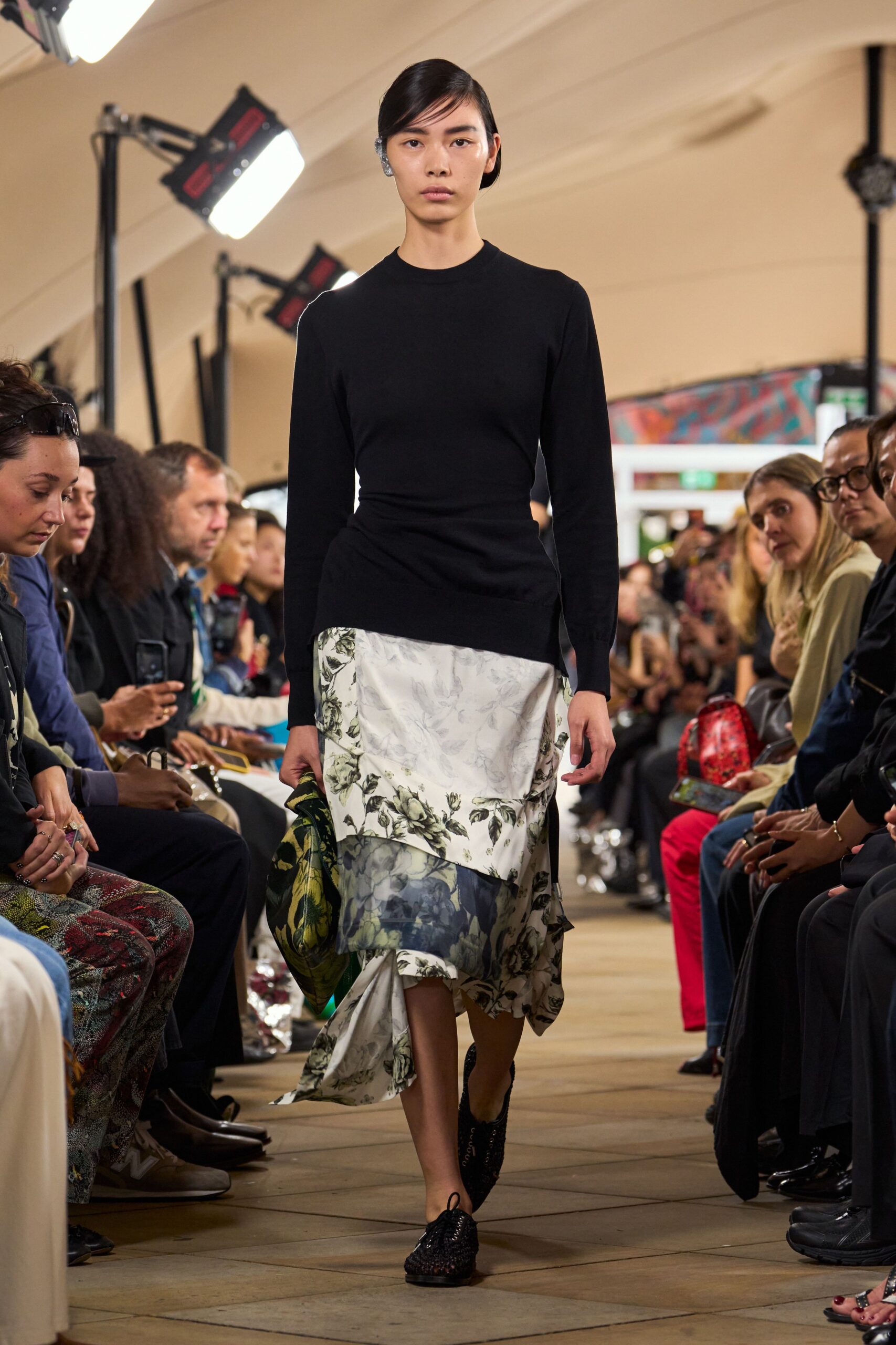 Black fine-knit top, drawn into a twisted drape at one hip. Asymmetric floral skirt spliced with cloudy mesh and a trailing handkerchief hem. Green floral pouch, slouched in one hand. Black woven lace-up flats. Crossing a conservatory after rain, gravel still stuck to the soles.