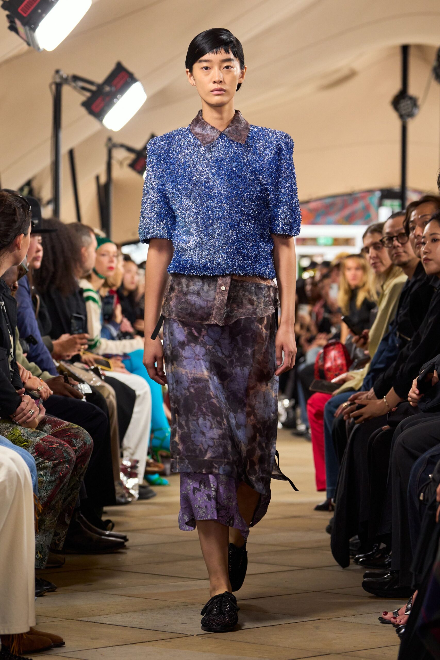 Cobalt tinsel-knit sweater with square shoulders and cropped short sleeves. Smoky floral sheer shirt, pointed collar spread over the neckline. Asymmetric midi skirt in the same blurred floral, hem dipping at one side. Black lace-up flats with a glittering woven vamp. Walking the long corridor to a wedding breakfast after rain.