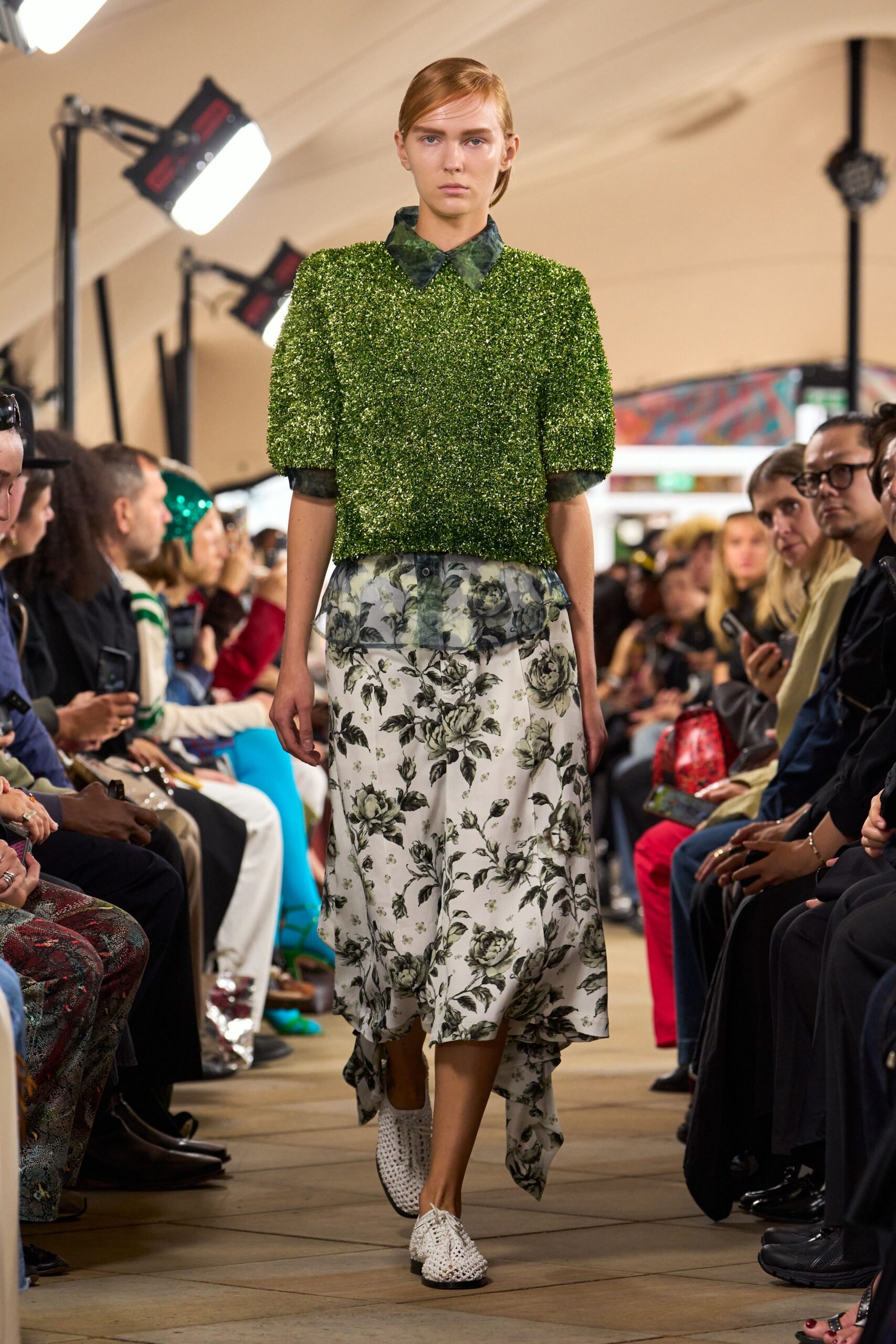 Moss-green tinsel knit top with rounded sleeves and a blunt cropped hem. Smoky green floral organza shirt, collar and cuffs left showing. Ivory skirt printed with charcoal roses, longer points dragging at the back. White woven leather oxfords with black-edged soles. Crossing a conservatory cafe just after the plant misting.