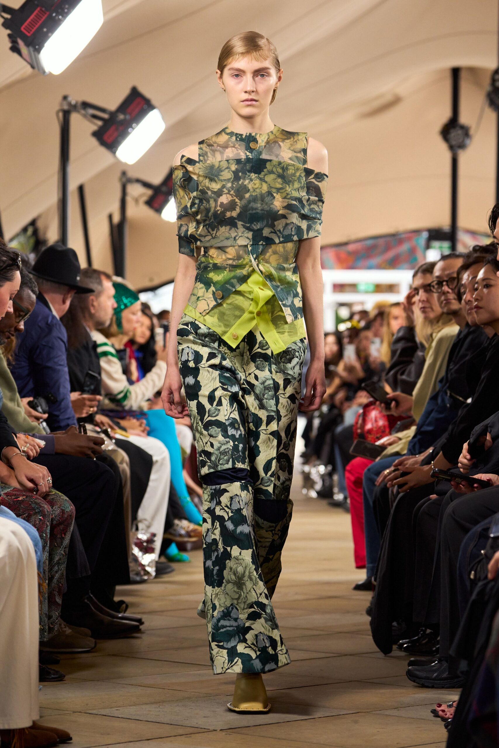 Sheer olive floral organza top, folded off the shoulders with one wooden button. Acid-lime poplin shirt peeking out, skewed placket crossing the waist. Wide floral trousers with dark inset knee bands and a long break. Olive square-toe boots. Under a marquee at noon, flowers crushed into the tablecloth.