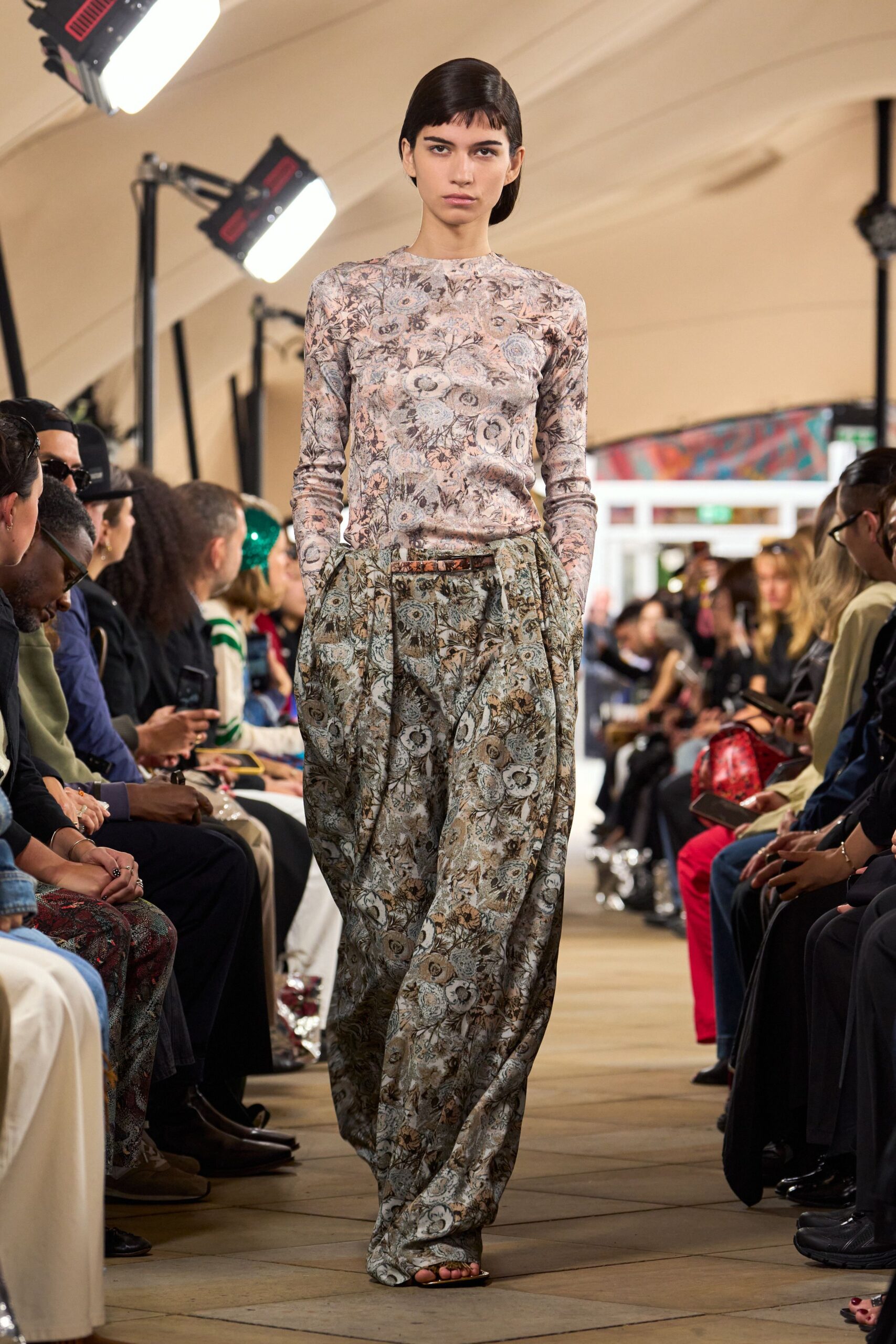 Second-skin floral knit in washed pinks, crewneck close to the collarbone. Loose floral trousers with deep front pleats and a pooled hem. Narrow brown belt, buckle nearly lost in the print. Flat black sandals, toes left bare. Walking the conservatory path after rain, gravel still dark underfoot.