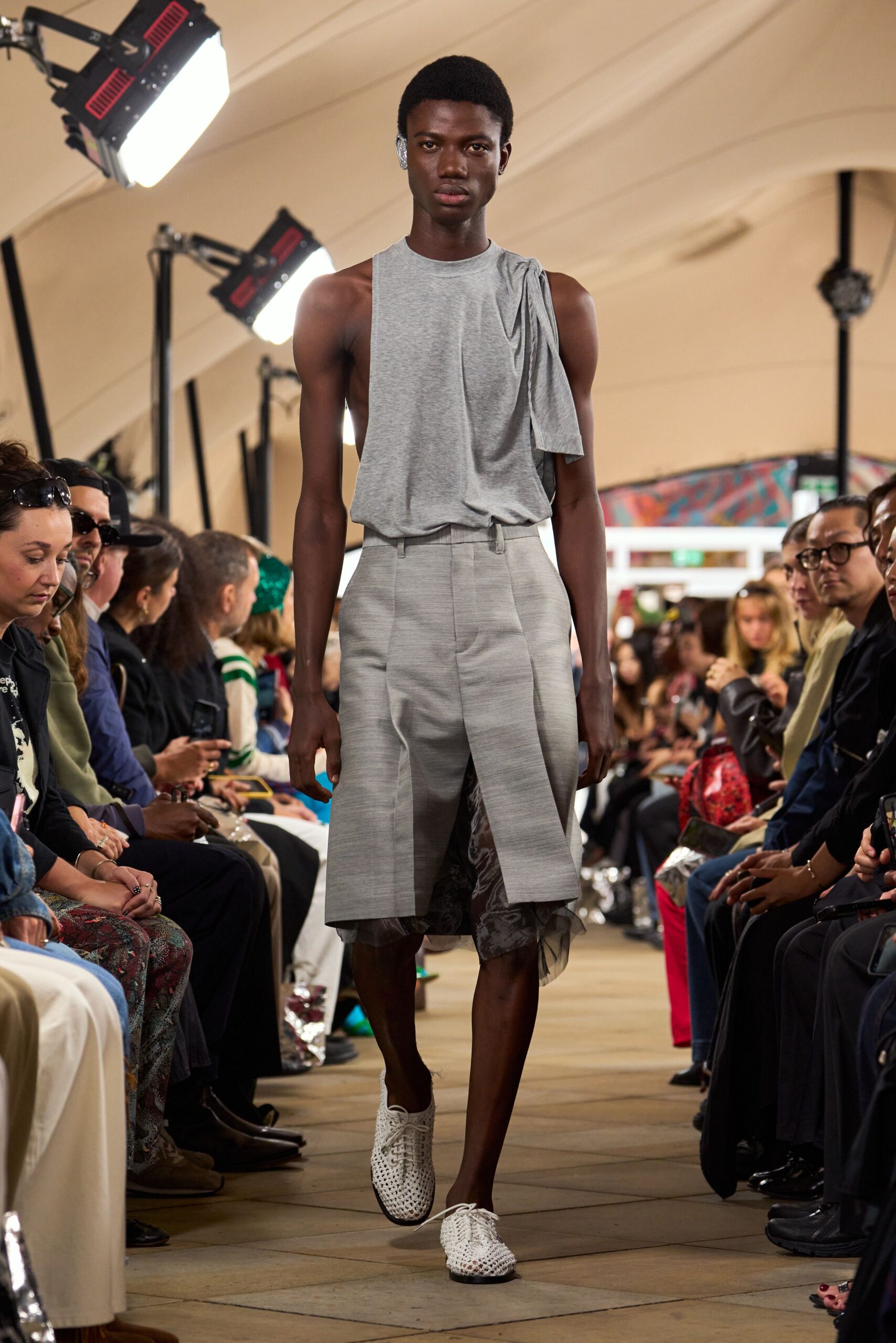 Heather-grey jersey top, one shoulder knotted into a hanging drape. Ash tailored culottes with a split apron panel and smoky sheer underlayer. White woven lace-up shoes, black sole peeking at the pointed toe. Walking through an airy civic hall after rain, paperwork still tucked under your arm.