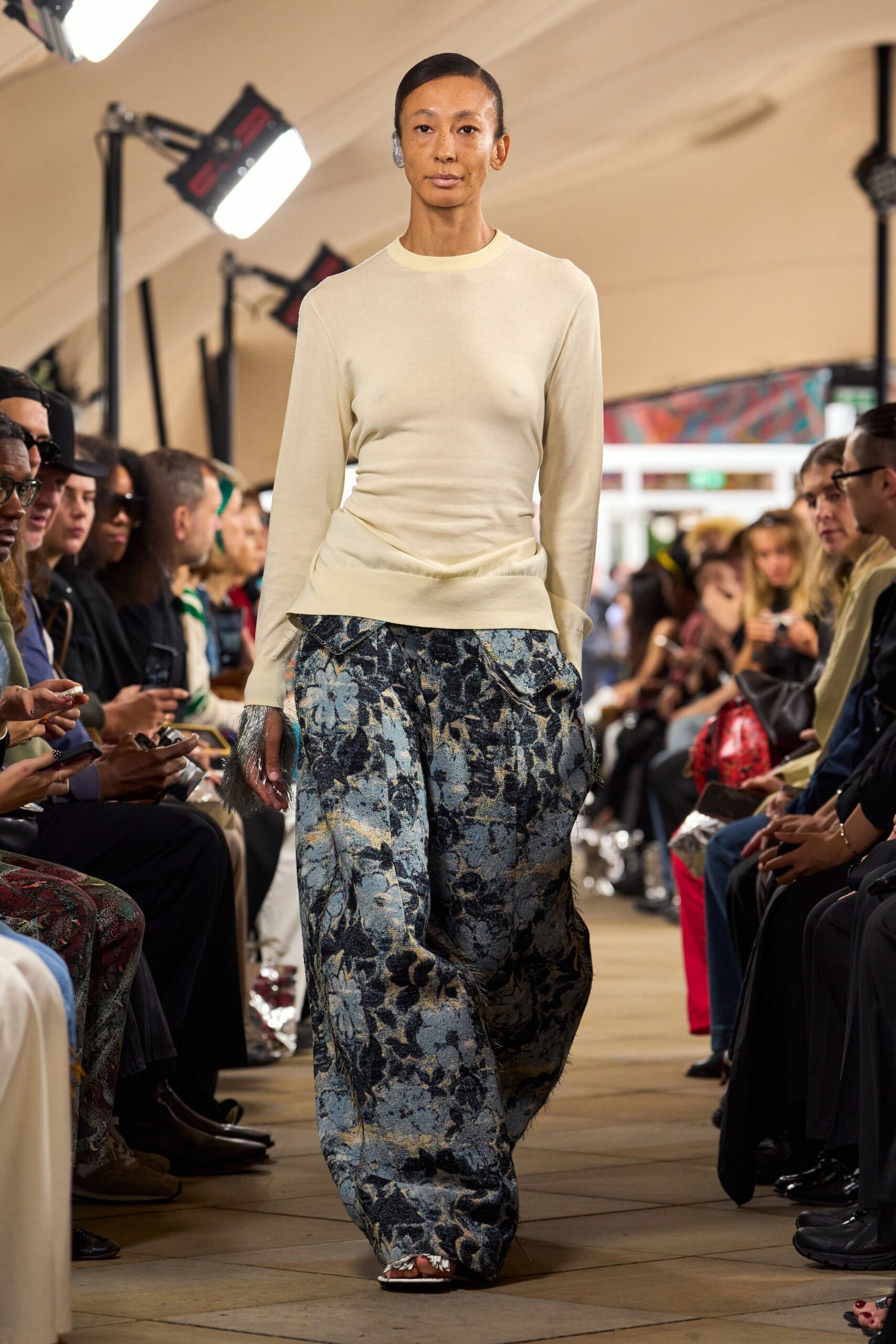 Cream fine-gauge knit top, ruched tight through the waist. Slate and powder-blue floral jacquard trousers, cut loose with fraying seams. Grey furry cuffs peeking from the sleeves. White sandals with shredded fringe over the toes. Walking into a hotel atrium after rain, hem already picking up grit.