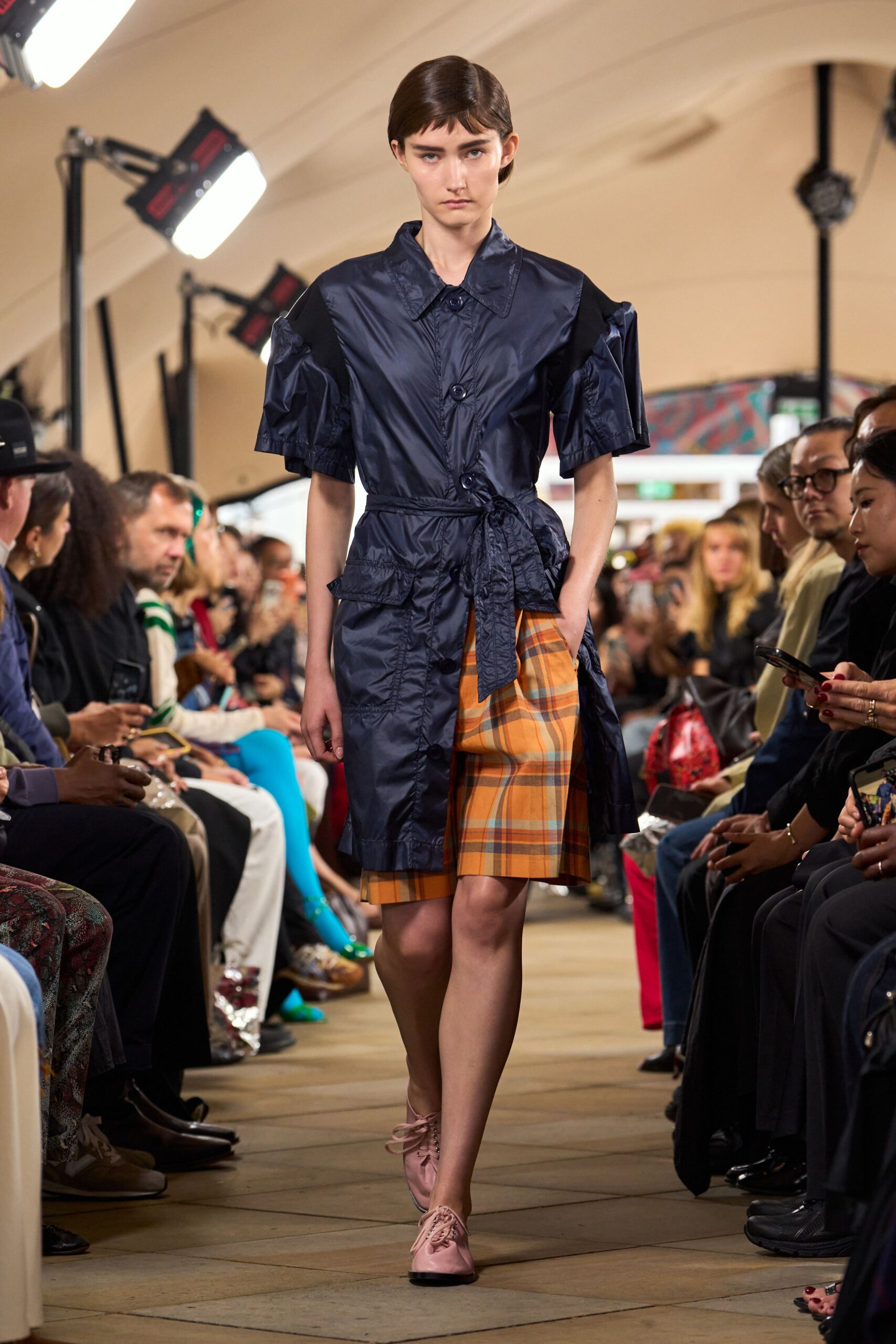 Inky nylon coat dress with puff sleeves and a long self-tie knot. Ochre plaid shorts peeking from the split front. Blush leather derbies with thin laces. Crossing the square after rain with shopping still under one arm.