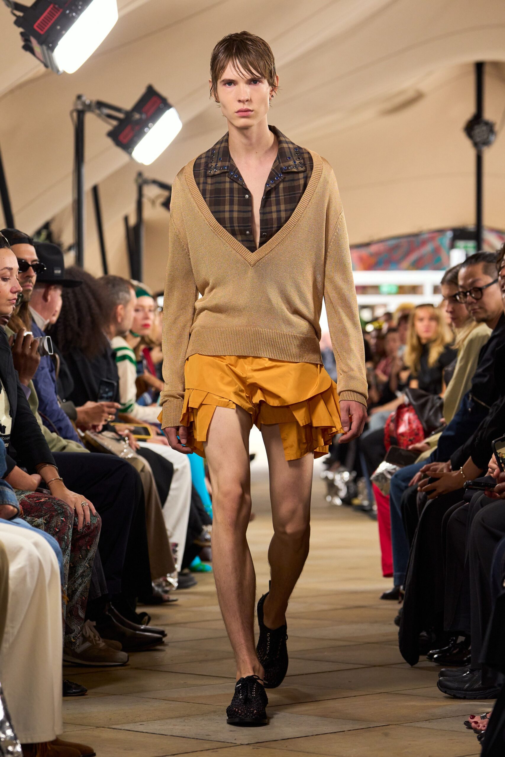 Brown tartan shirt, collar and placket studded with blue grommets. Deep-V camel knit sweater, ribbed trim hanging loose at the hip. Marigold poplin shorts with split ruffle panels flicking below the hem. Black open-weave lace-up shoes. Walking out of a matinee into warm wind and traffic grit.
