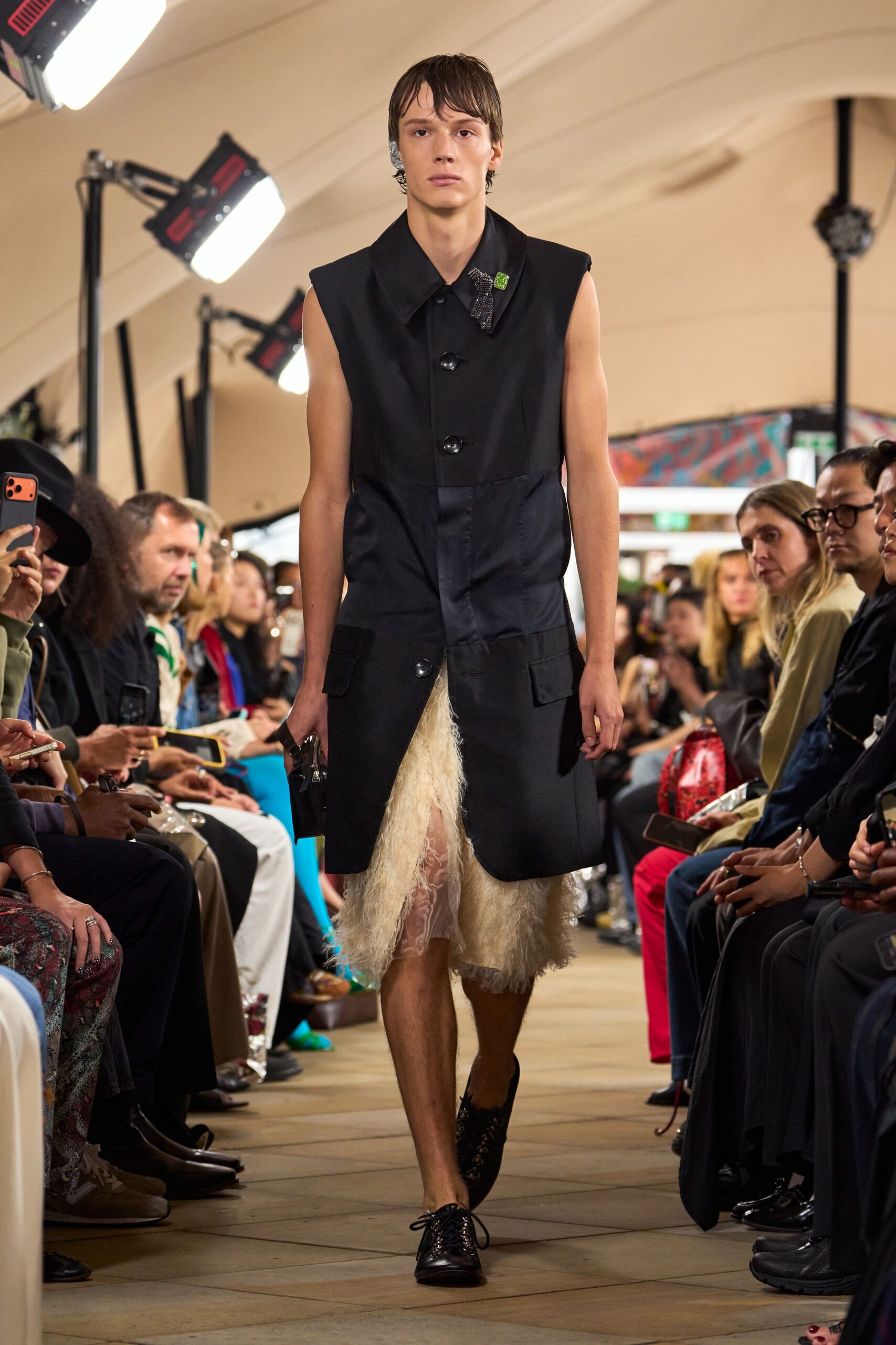 Black sleeveless coat with oversized collar and three glossy horn buttons. Green square pin beside a beaded silver insect brooch. Cream lace skirt veiled in shaggy mohair strands. Black lace-up shoes, low and narrow at the toe. Crossing a drafty town hall after someone else's wedding ran long.