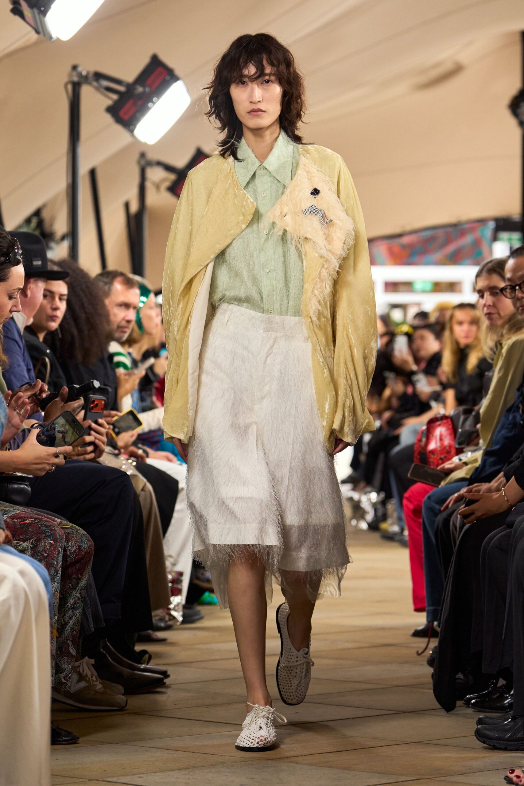 Pale mint sheer blouse with a long pointed collar. Butter velvet jacket cut open at the chest with shaggy cream trim. White knee skirt with a cloudy sheer overskirt hanging uneven at the hem. White woven lace-up shoes with black-edged soles. Crossing a conservatory corridor while the sprinklers tick on at dusk.