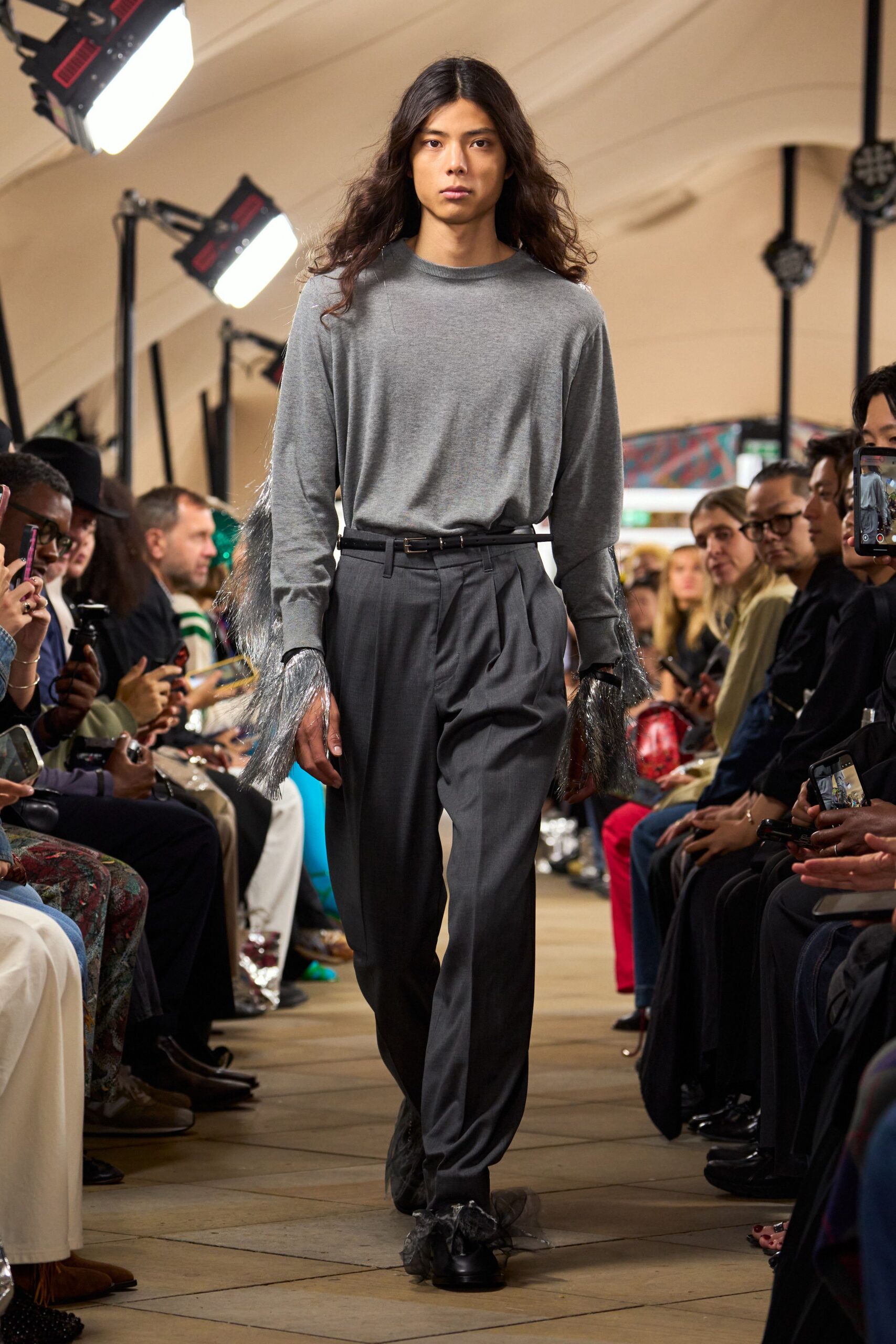 Heather-grey fine knit sweater with silver fringe bursting from the cuffs. Narrow black belt with a small brass buckle. Charcoal wool trousers, double-pleated and cut loose through the leg. Black shoes muffled by wisps of sheer black tulle. Crossing a museum atrium after someone dropped glitter in the coat check.