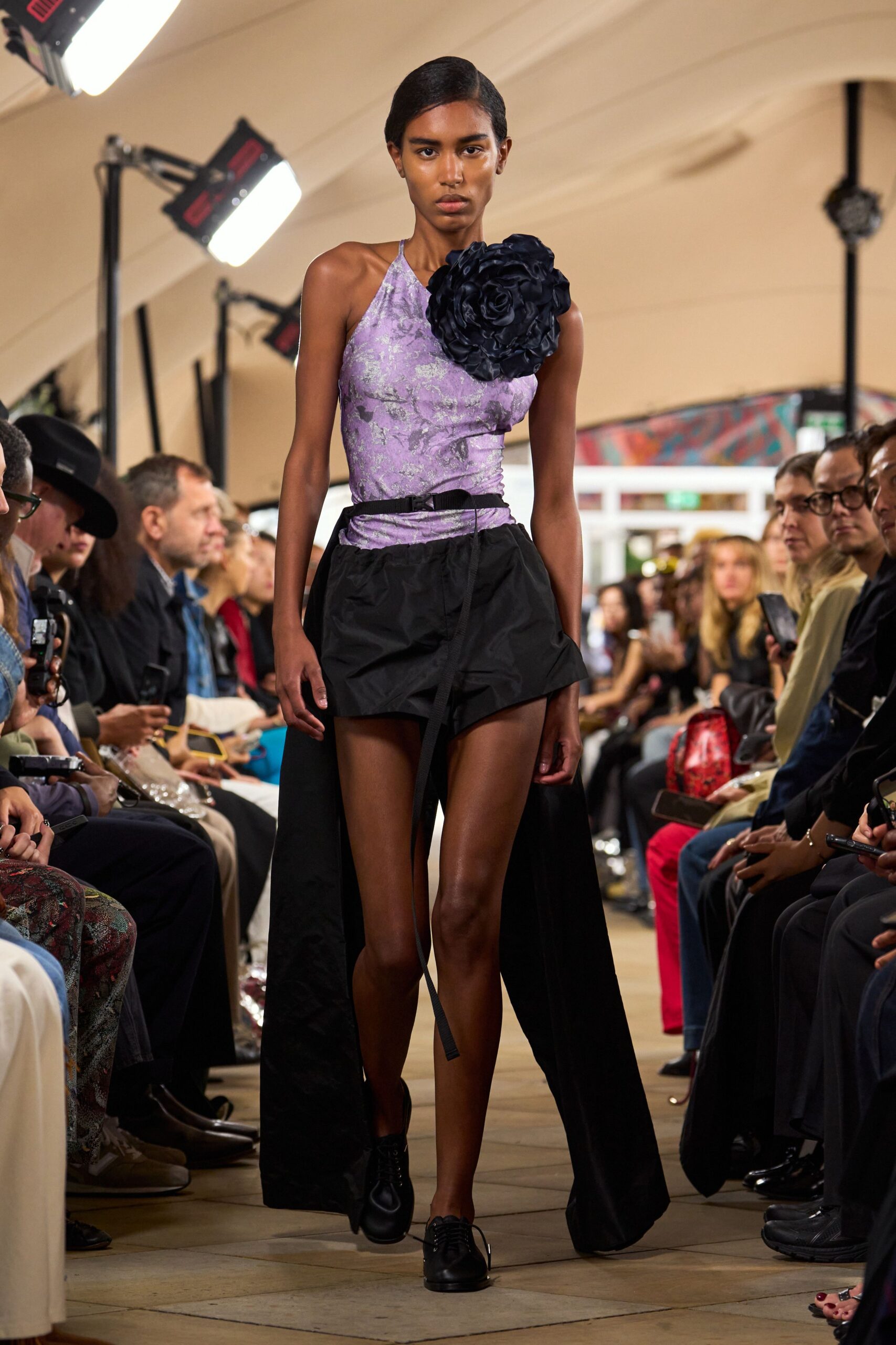 Lilac foil-printed halter bodysuit, one shoulder swallowed by a black rosette. Black nylon micro shorts with a floor-length overskirt split open behind. Webbing belt with one strap end hanging past the knee. Black leather derbies, blunt and compact. For crossing a hotel lobby after midnight with flowers already crushed.