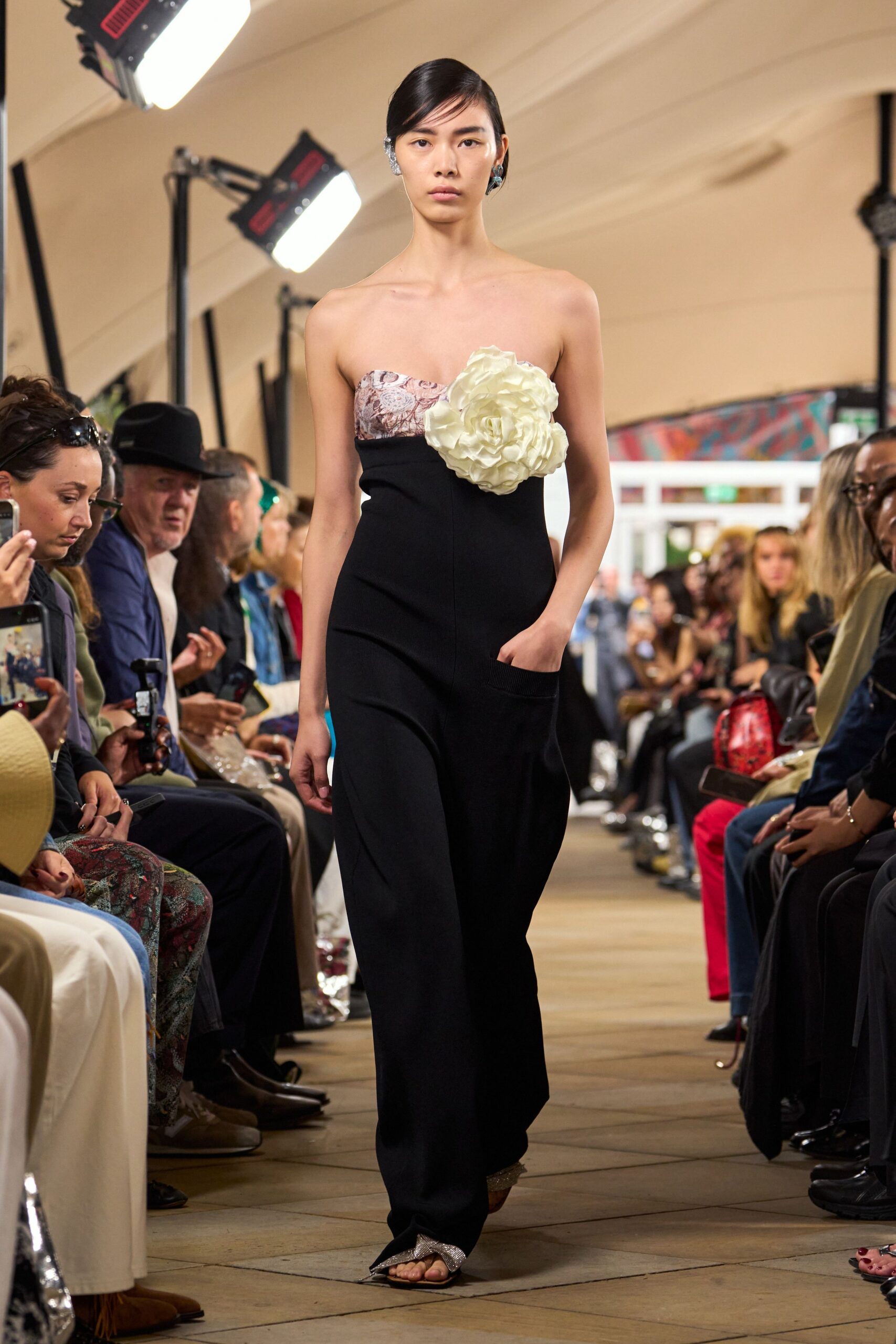 Black strapless jumpsuit with a mauve toile bust and deep patch pocket. Oversized ivory rosette pinned high across one side of the chest. Blue crystal ear clips. Silver glitter sandals peeking from the pooling hem. Walking into a wedding lunch after changing out of the bridesmaid dress.
