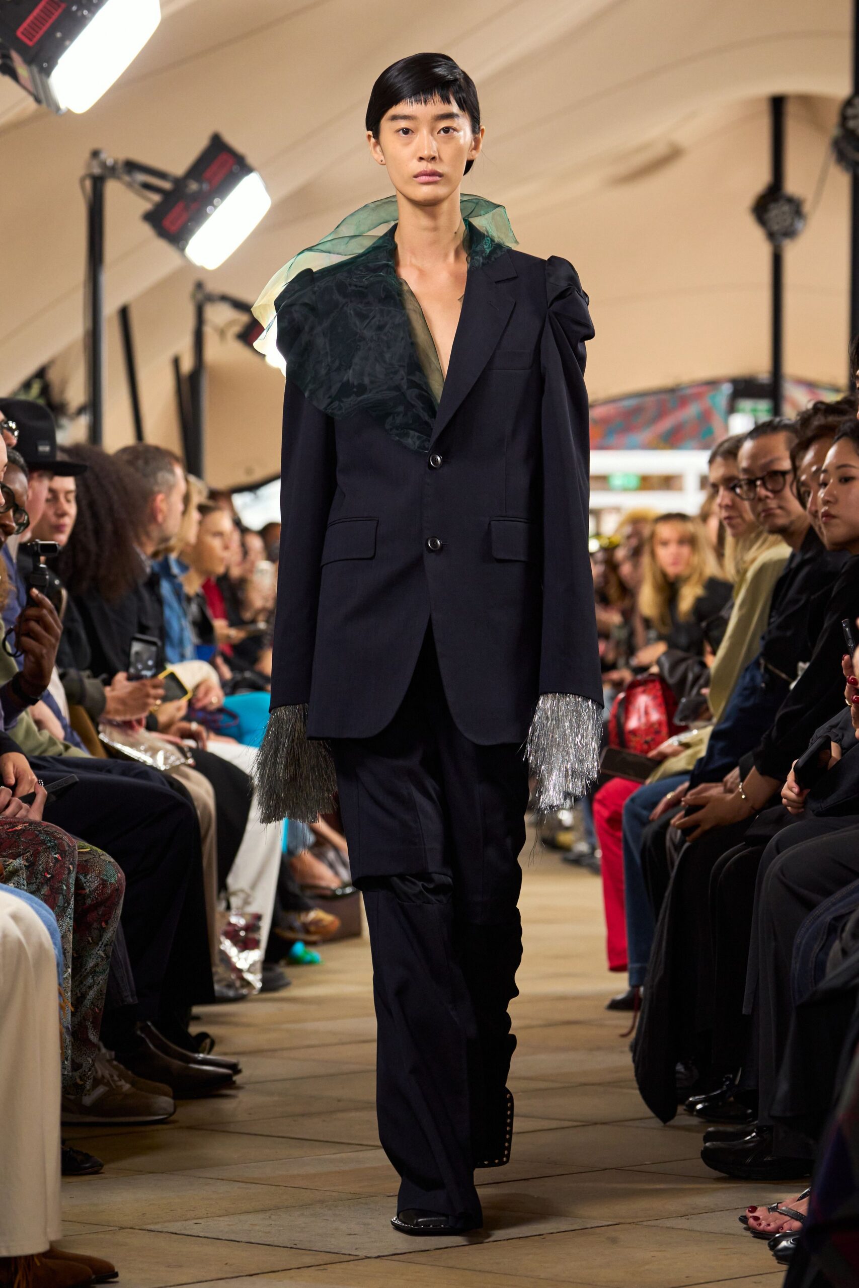 Sheer green organza scarf blown wide across one shoulder. Inky tailored blazer with a gathered puff shoulder and silver fringe cuffs. Matching trousers, slit open at one knee and puddling slightly. Square-toe black shoes. Crossing the lobby after the orchestra packed up, tinsel still stuck to your sleeves.