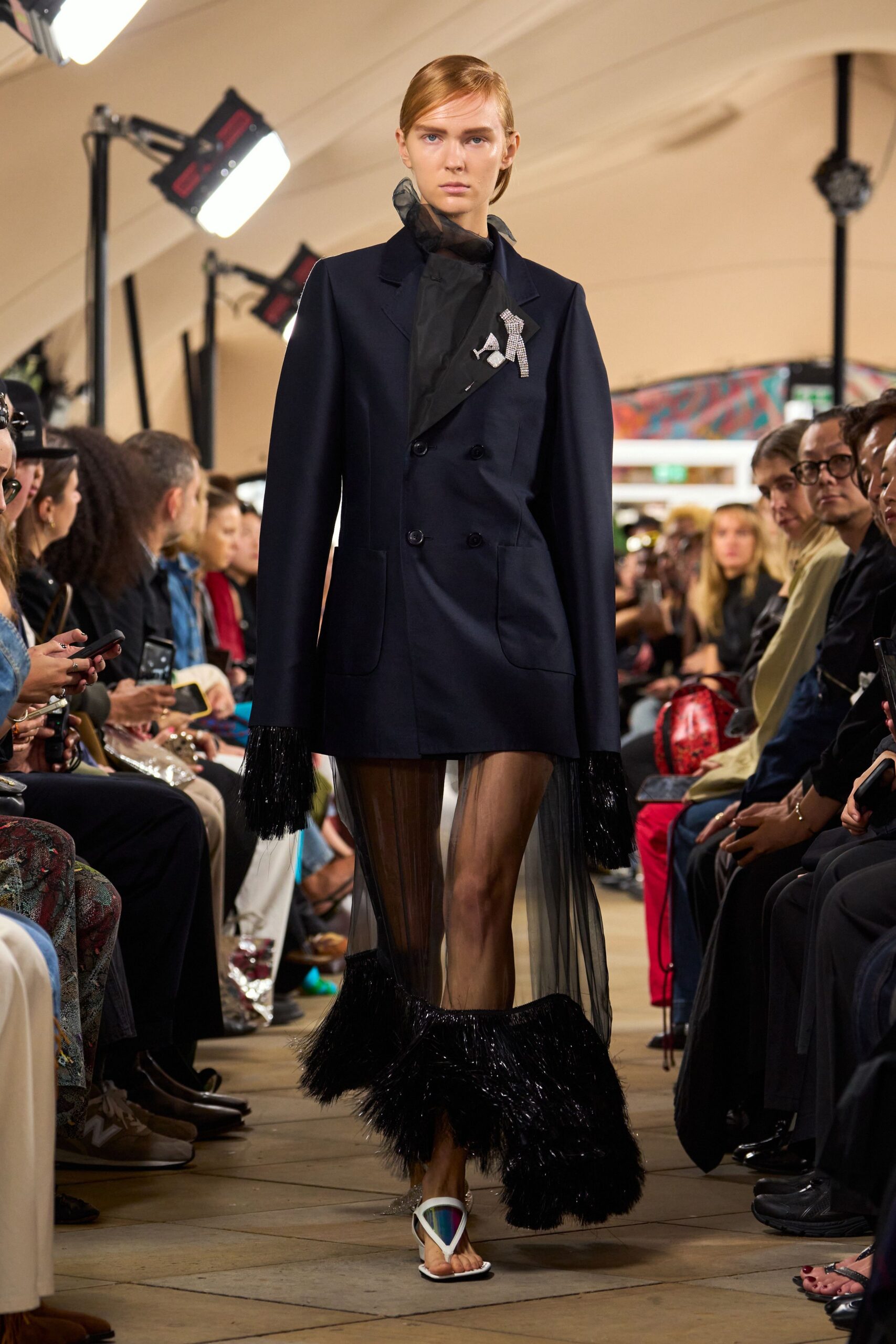 Ink double-breasted blazer with rounded patch pockets and a rhinestone bow brooch. Black organza standing up in a loose transparent collar. Sheer black chiffon falling from the hips to fringed bands. White thong sandals with iridescent shield vamps. Crossing the hotel atrium after a benefit, still wearing the corsage.