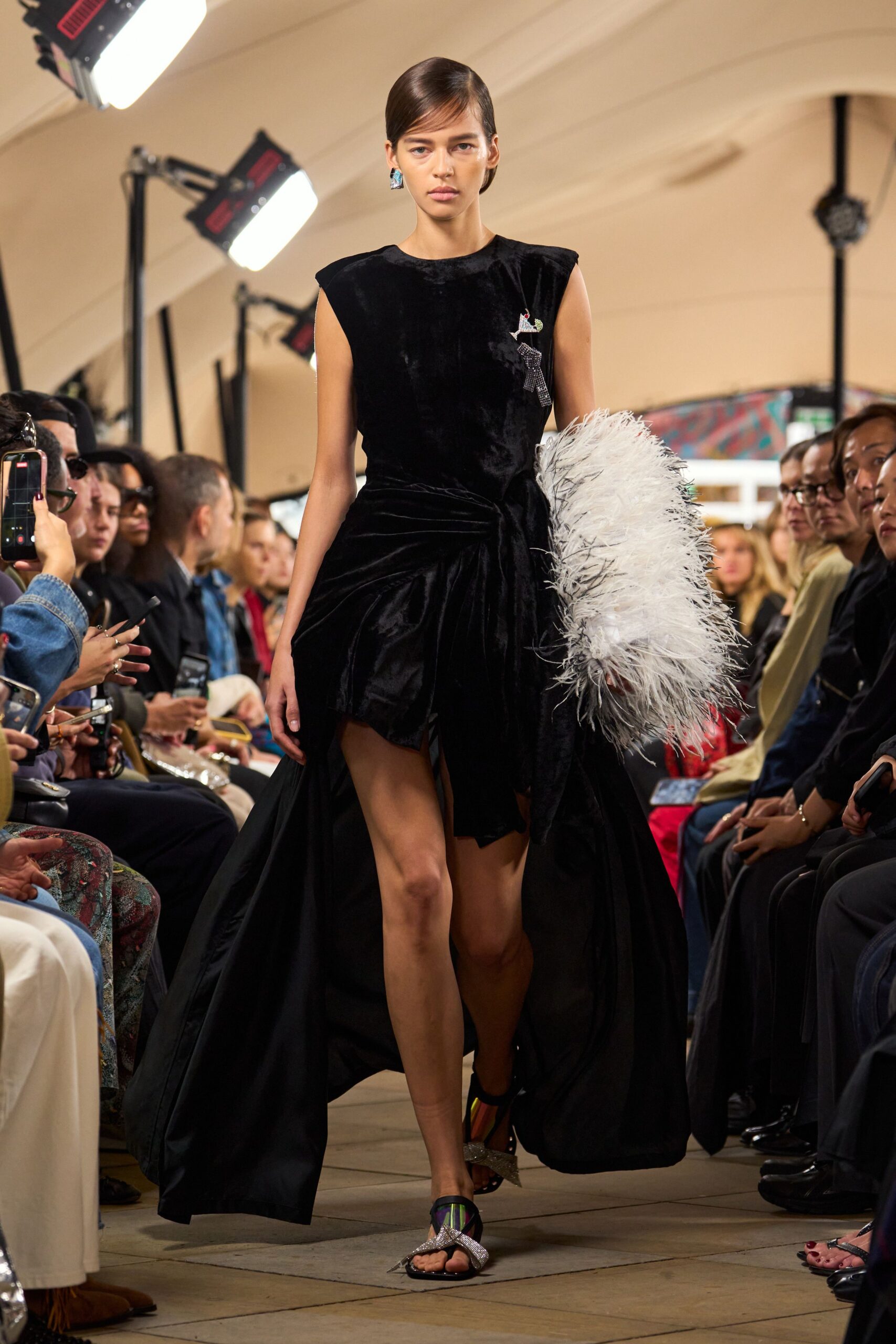 Black velvet dress with padded cap shoulders and a draped train. Rhinestone donkey pinned high on the chest. White feather stole crushed under one arm. Crystal crossover straps over iridescent insoles. Crossing a hotel ballroom after the auction, still carrying the centerpiece.
