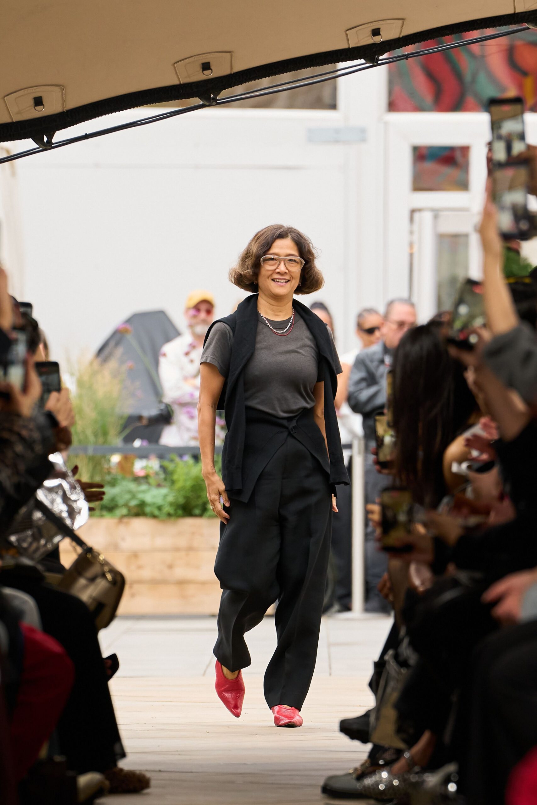 Clear acetate frames with a squared-off brow. Washed charcoal jersey tee with softly rolled sleeves. Black knit draped over the shoulders, sleeves hanging loose. Black wrap-front trousers with a folded crossover waist. Tomato red leather flats, creased across the vamp. Walking the garden aisle while everyone lifts a phone.