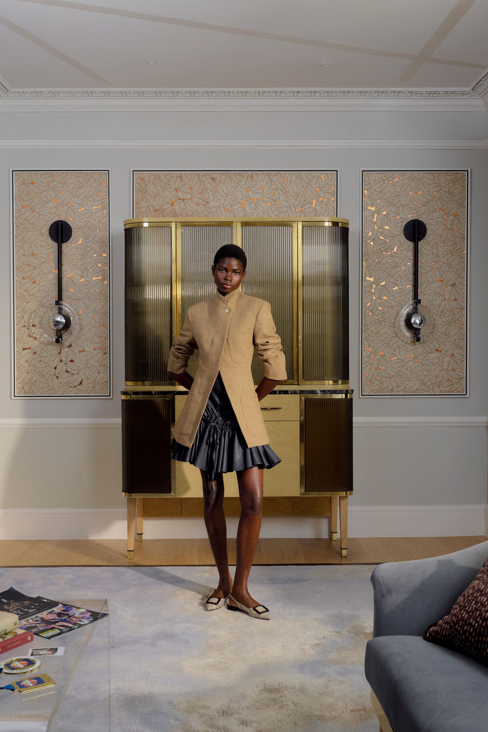 Camel tailored jacket with a high stand collar and curved split front. Black satin mini, dropped waist gathered by a drawstring into a flounced hem. Cream slingback flats with oversized square buckles. Hotel suite carpet, deciding whether to go downstairs for one more drink.