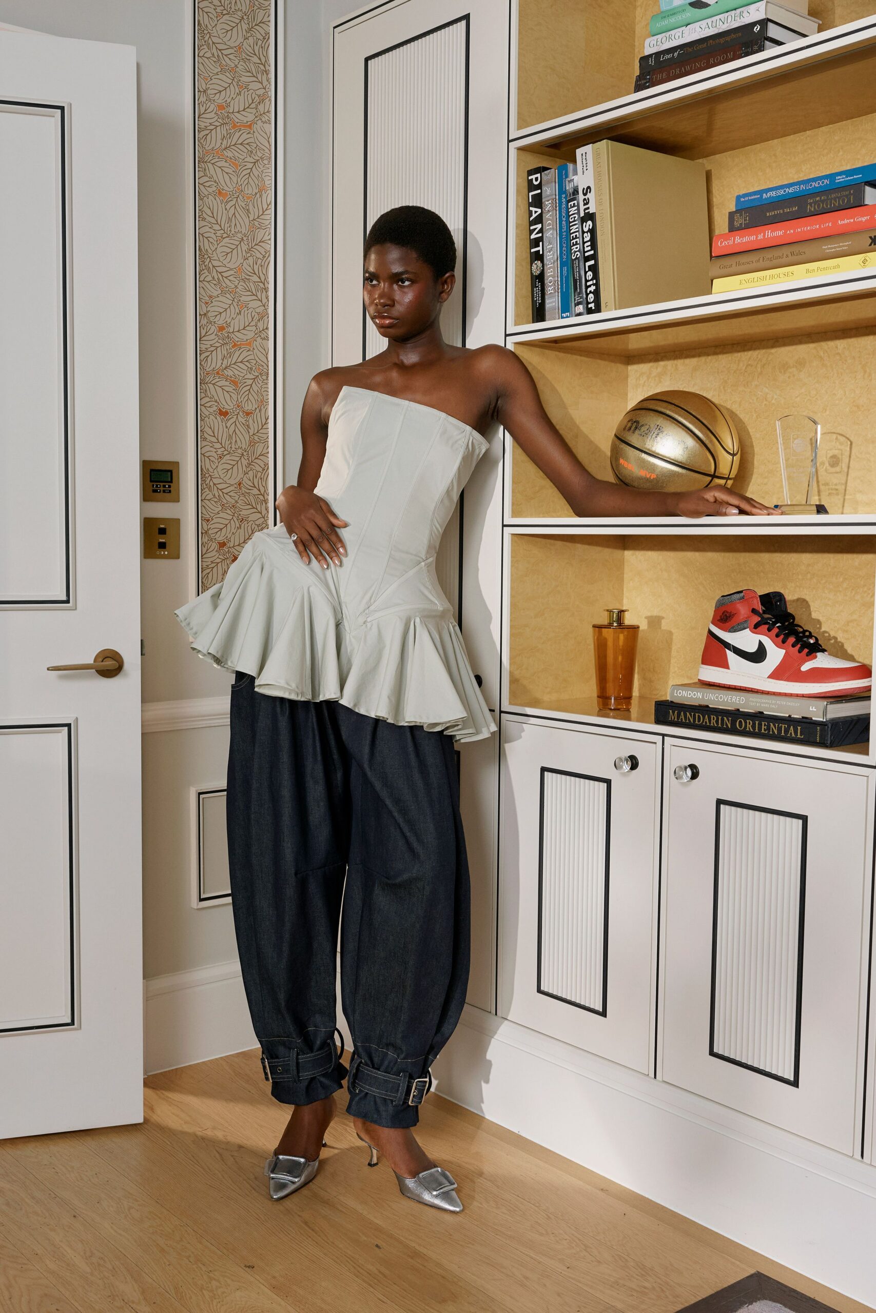 Pale dove strapless peplum top, seamed like a corset and cut uneven. Dark indigo denim barrel trousers, buckled tabs cinching both cuffs. Silver pointed slingbacks with oversized square buckles. Leaning on the built-ins before dinner, trophy shelf at your elbow.
