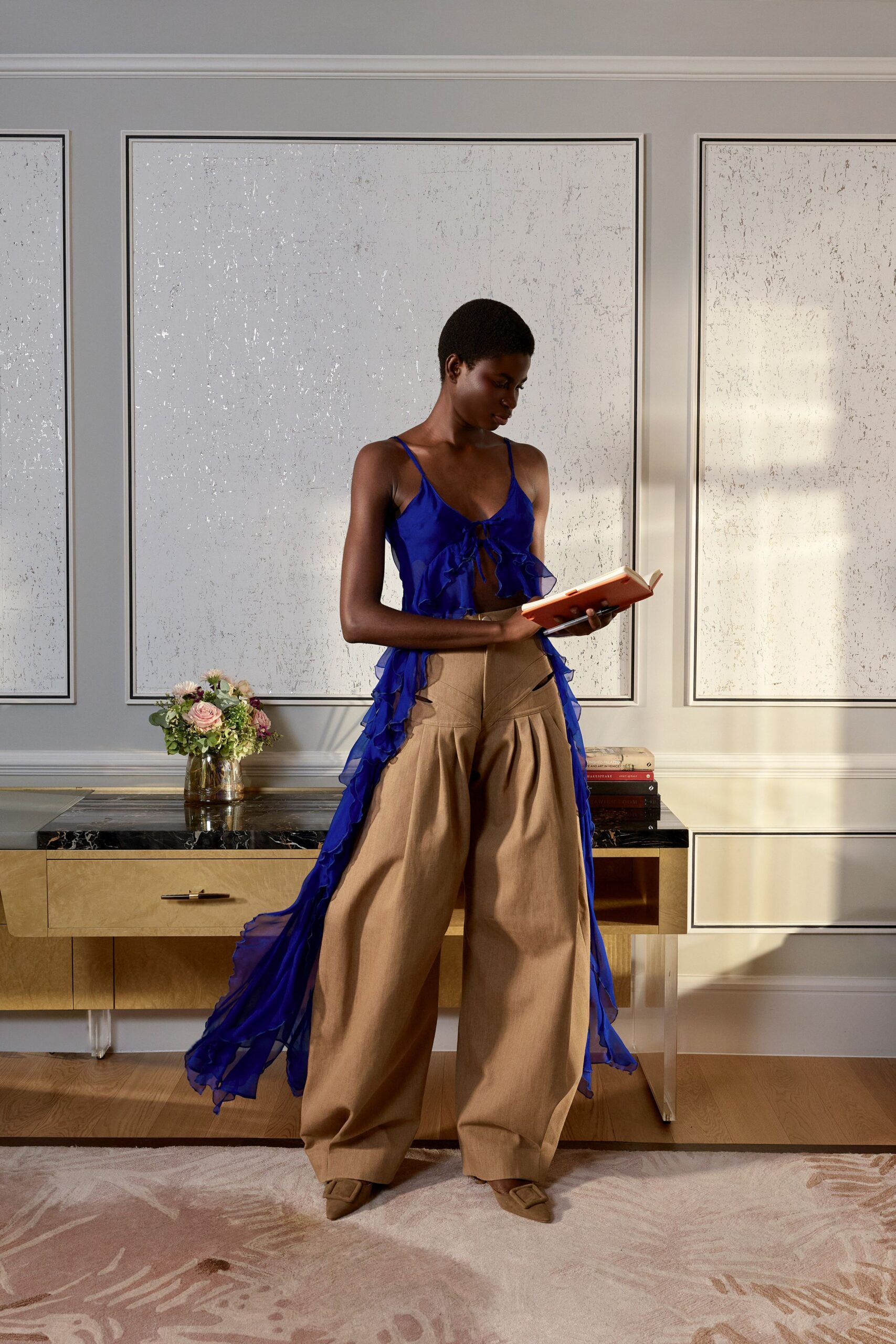 Cobalt chiffon camisole with a tied front and ruffled high-low train. Camel wide-leg trousers with deep front pleats and slashed hip openings. Matching pointed mules with covered square buckles. Late sun in a borrowed apartment, reading before anyone else arrives.