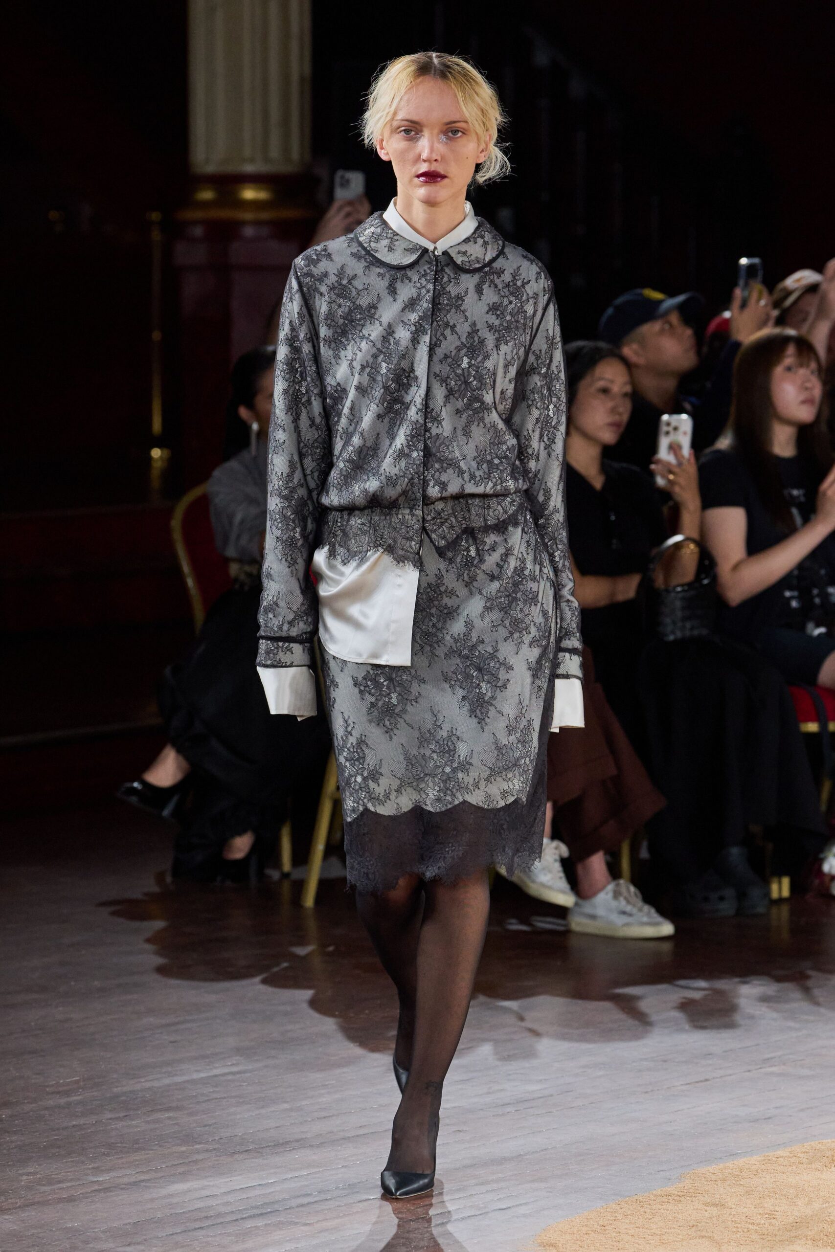 Smoke satin blouse under black floral lace, rounded collar edged in piping. Matching lace skirt over pale slip, scalloped hem dropping into darker lace. Sheer black hosiery. Black pumps, squared toe. Leaving a courthouse wedding after the flowers have already wilted.