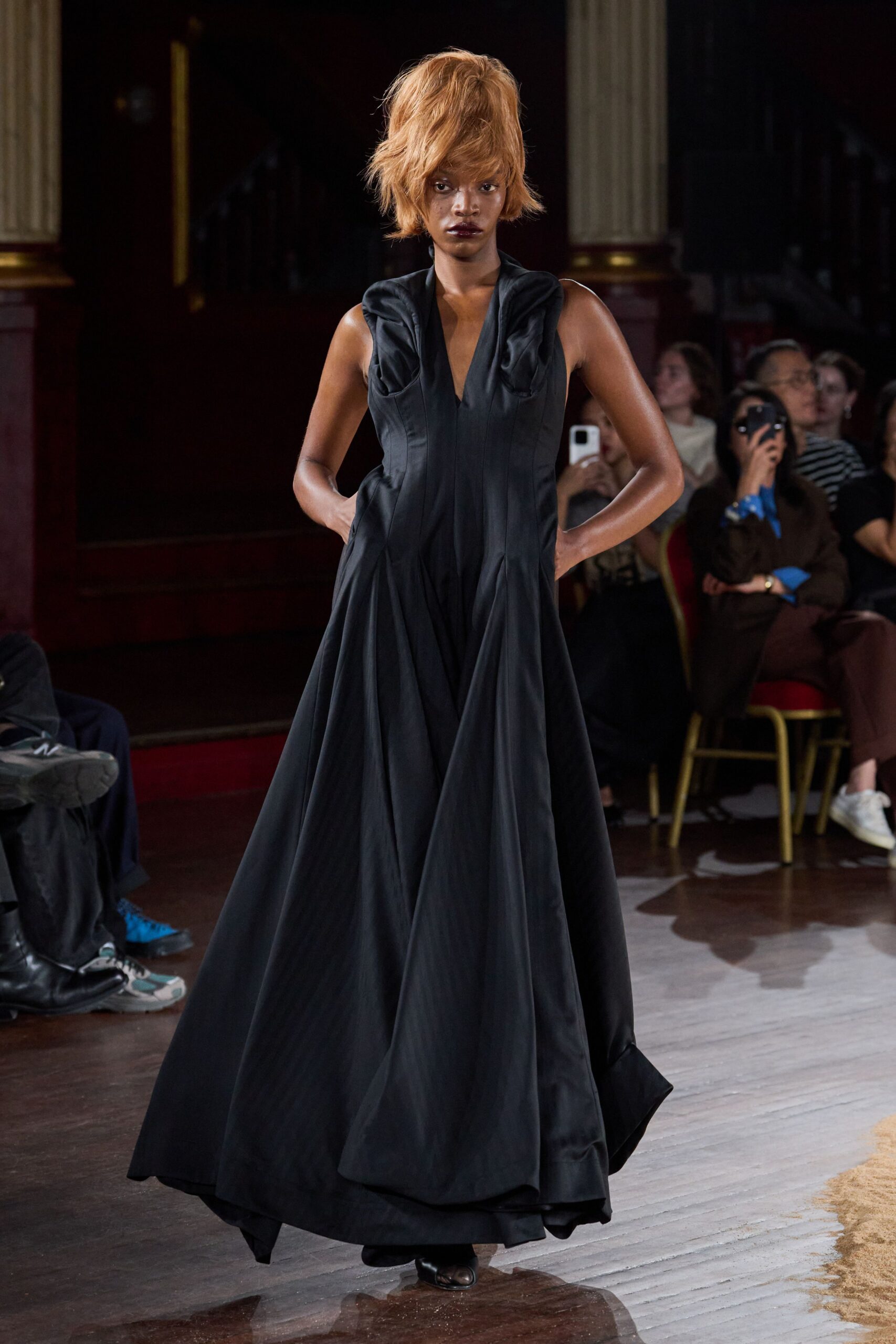 Black satin floor-length gown, sculpted shoulder folds and deep V neck. Black square-toe shoes just breaking the hem. Crossing a municipal ballroom floor after the orchestra has packed up.