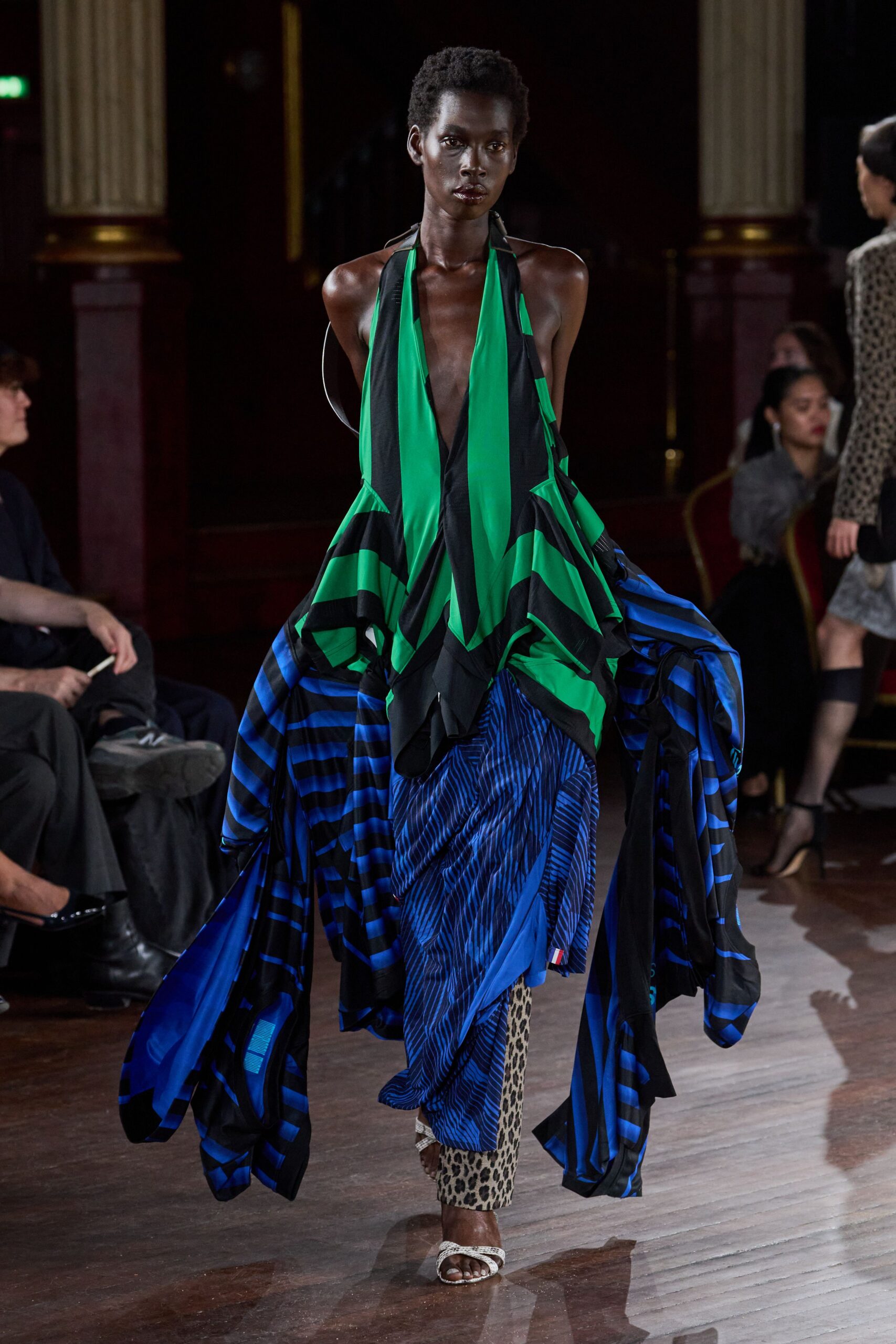 Plunging halter dress in emerald and black jersey, draped into long trailing panels. Electric blue striped trousers, softly pleated and tapering to leopard-print calves. White braided mules with a square open toe. Crossing a hotel ballroom after midnight, carrying half the curtains with you.