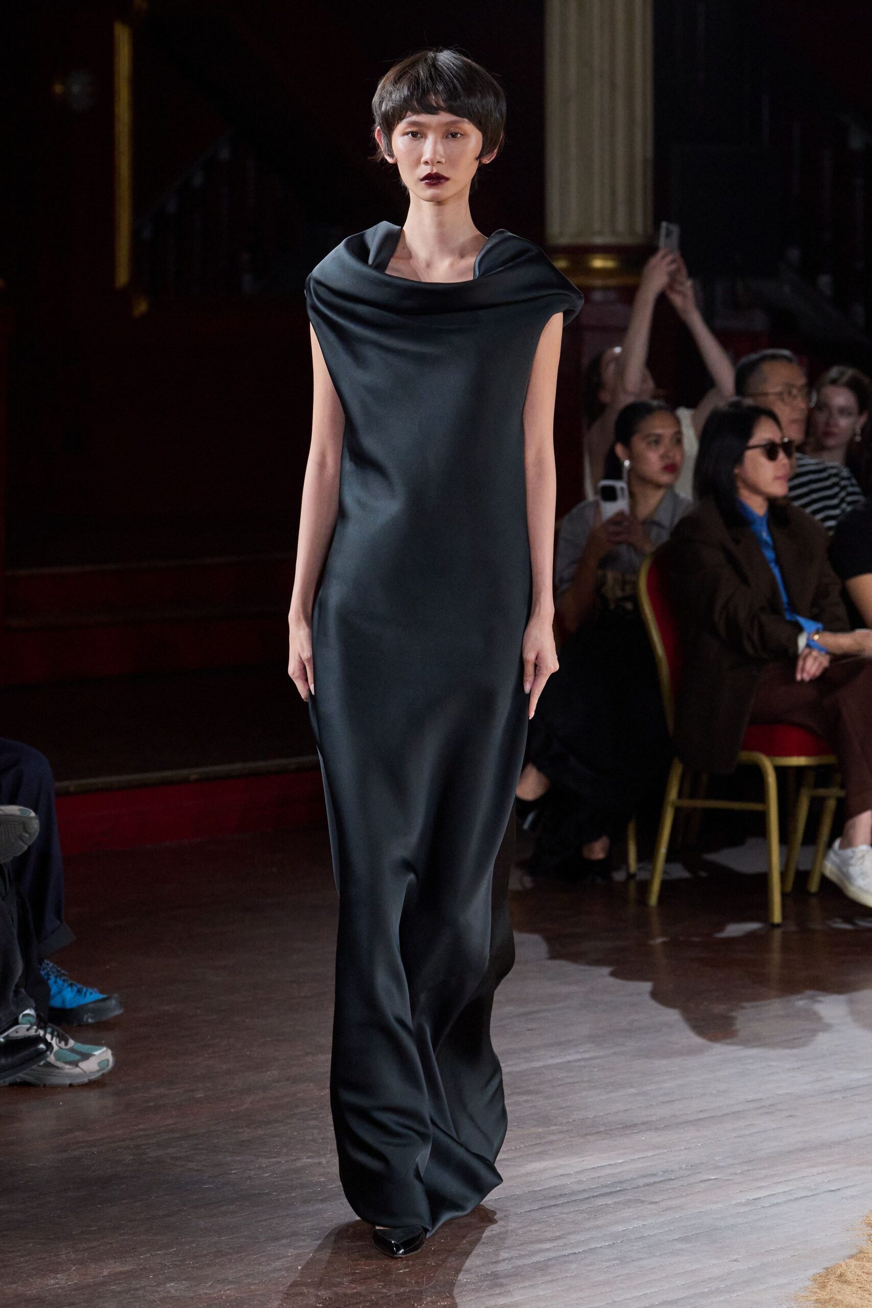 Black satin column gown with a wide folded bateau collar. Pointed black shoes barely breaking the hem. Crossing a hotel ballroom after midnight, carrying yourself quieter than everyone else.