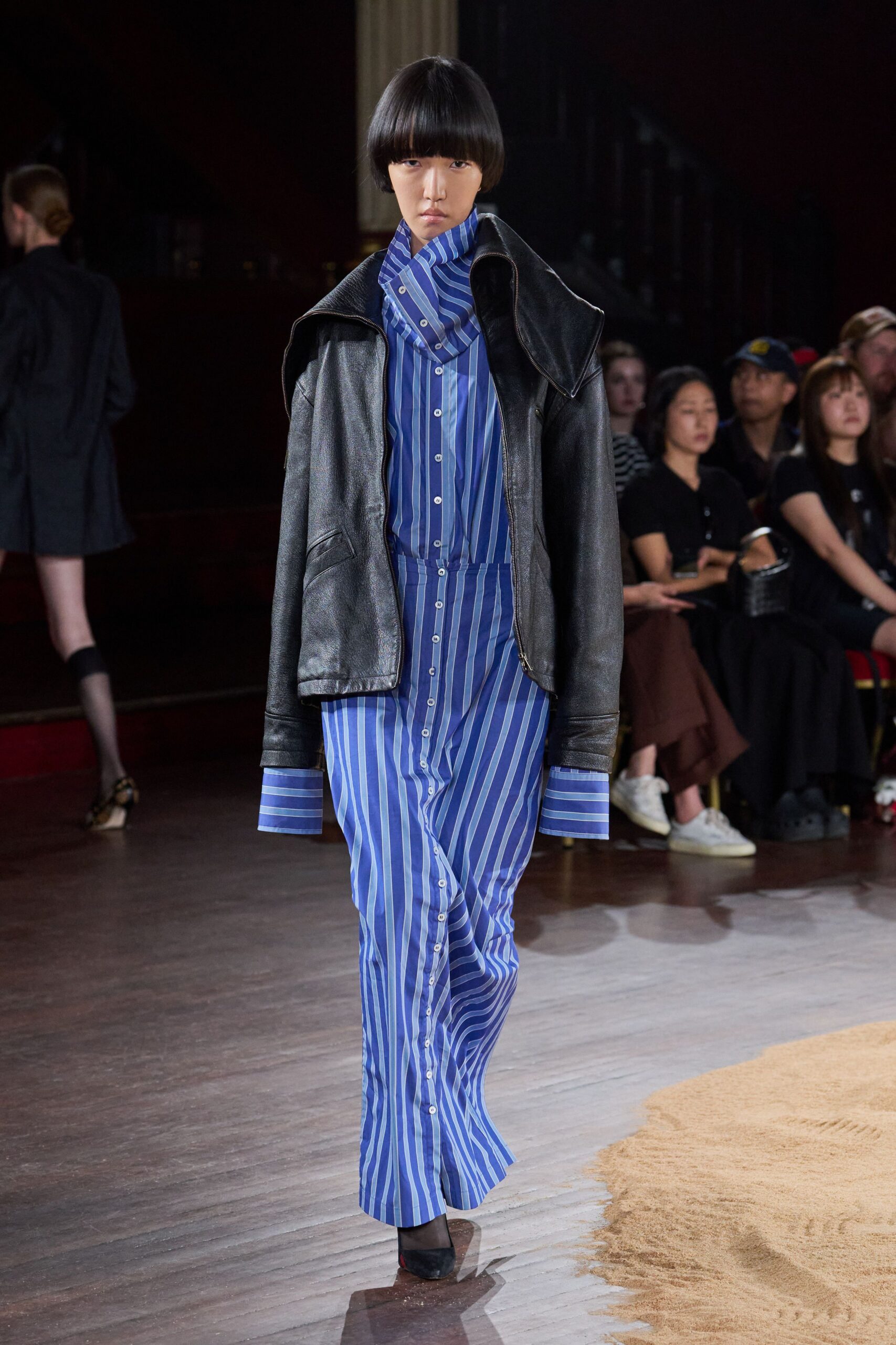 Black leather jacket with an oversized collar folded into wide wings. Blue striped shirting cut long and straight, buttons running to the hem. Matching striped panel looped high at the throat. Black pumps with a flash of red sole. Walking into a town-hall meeting after sleeping in yesterday's office clothes.