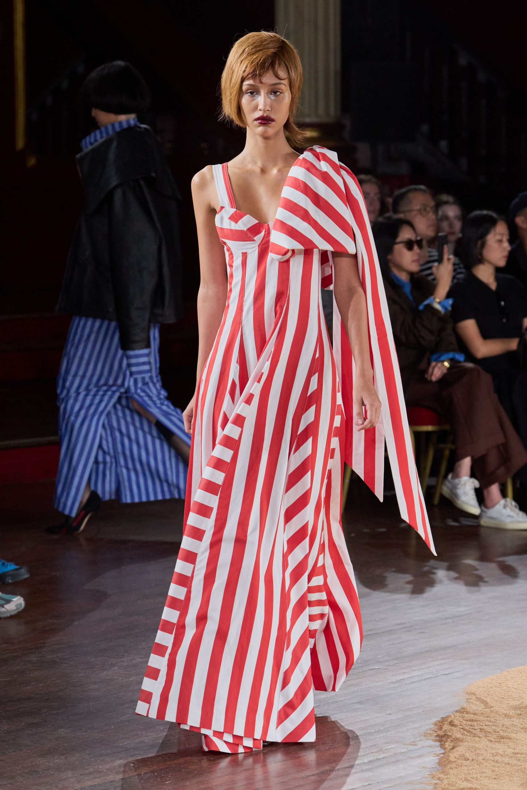 Red-and-white striped satin gown, one shoulder exploding into a long bowed sash. White shoulder strap left plainly visible above the sweetheart bodice. Walking down a pier with carnival canvas snapping in the wind.