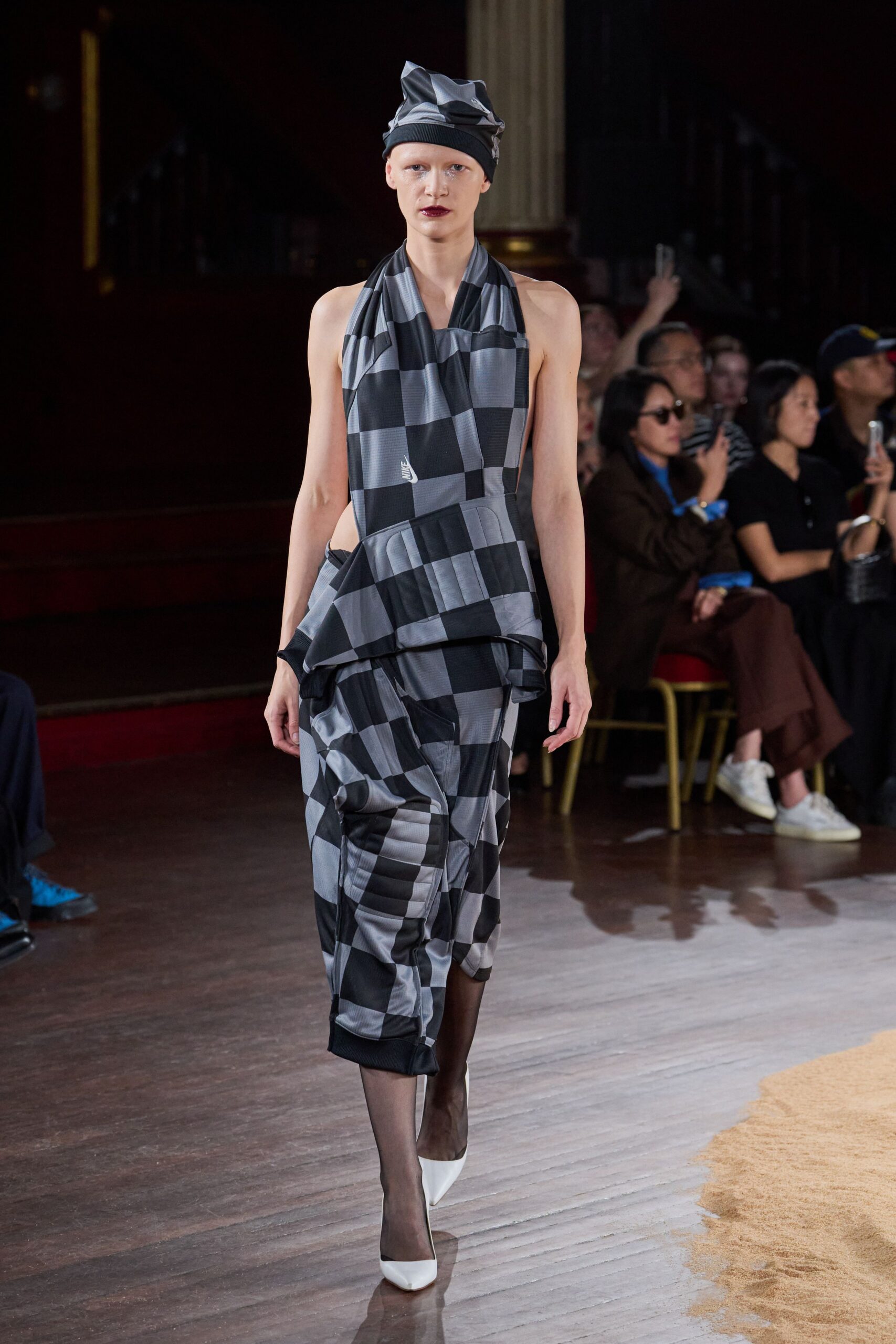 Twisted grey-and-black checked jersey knotted into a soft peak. Draped checked knit halter, open at the sides with a folded peplum. Drop-crotch checked trousers ruched into ribbed cuffs at the calf. Smoke sheer tights. White pumps with a hard pointed toe. Crossing a varnished town-hall floor after rain, carrying gossip upstairs.
