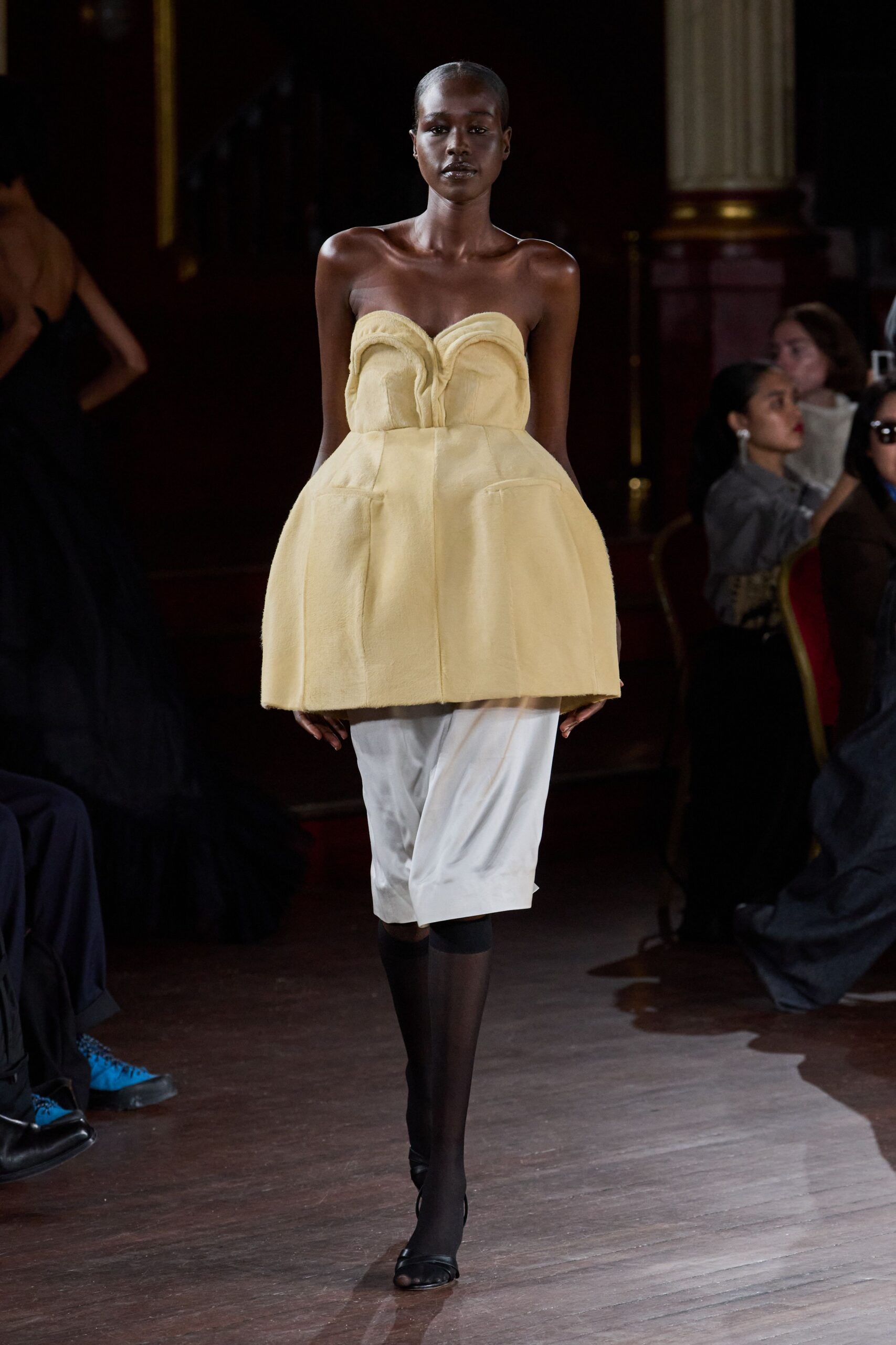 Buttercream strapless dress with sculpted sweetheart folds and a stiff bell-shaped overskirt. White satin pencil underlayer, wrinkling softly beneath the outer shell. Black sheer stockings ending mid-calf. Black peep-toe slingbacks. Walking through a municipal ballroom after the flower arrangements have started to wilt.