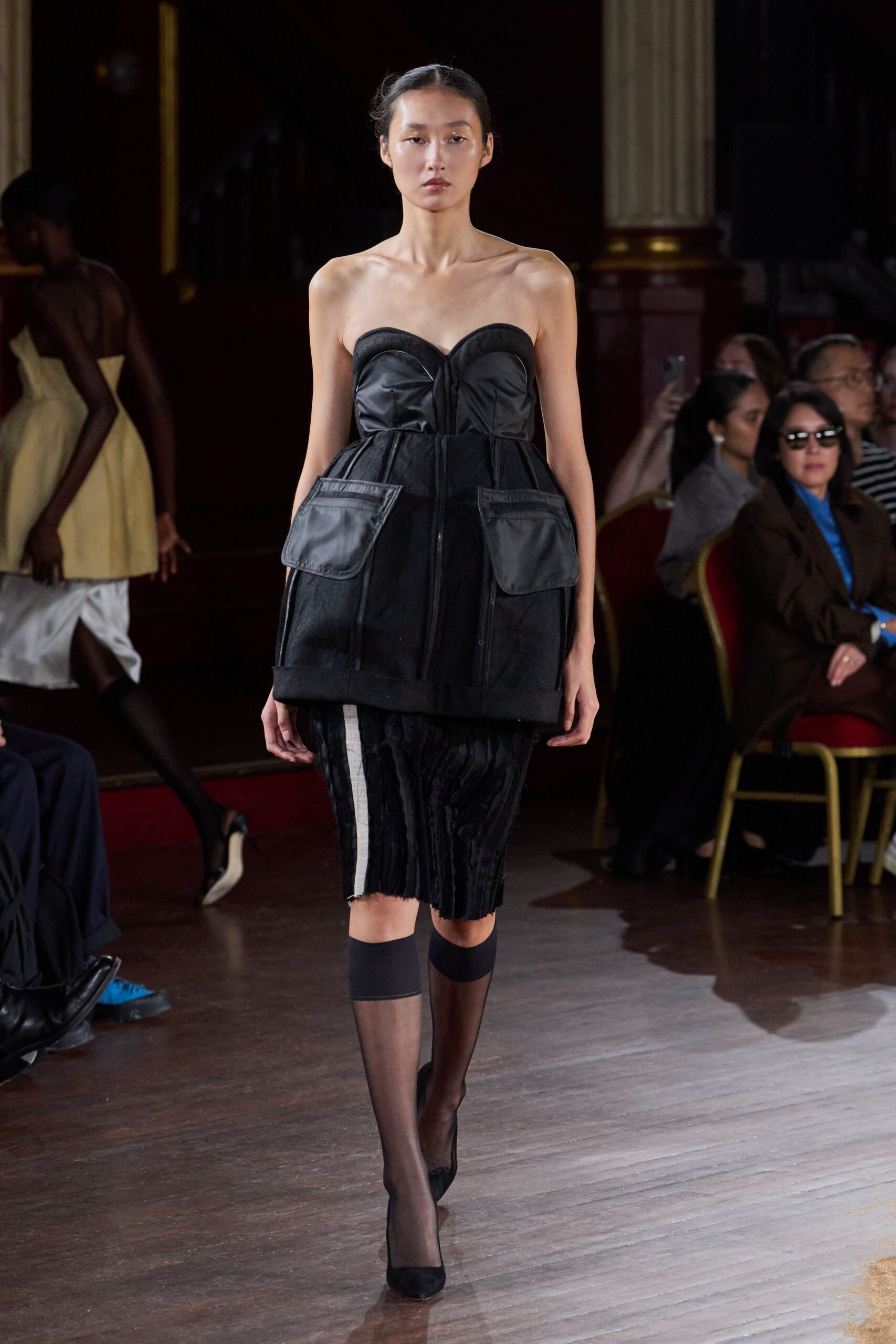 Black strapless bustier dress with satin cups and bulging cargo pockets. Black knee skirt spliced with narrow pleats and one chalk side panel. Sheer black stockings ending in opaque bands below the knee. Black suede pumps with a blunt almond toe. Walking through a municipal hall after midnight, carrying nothing but your keys.