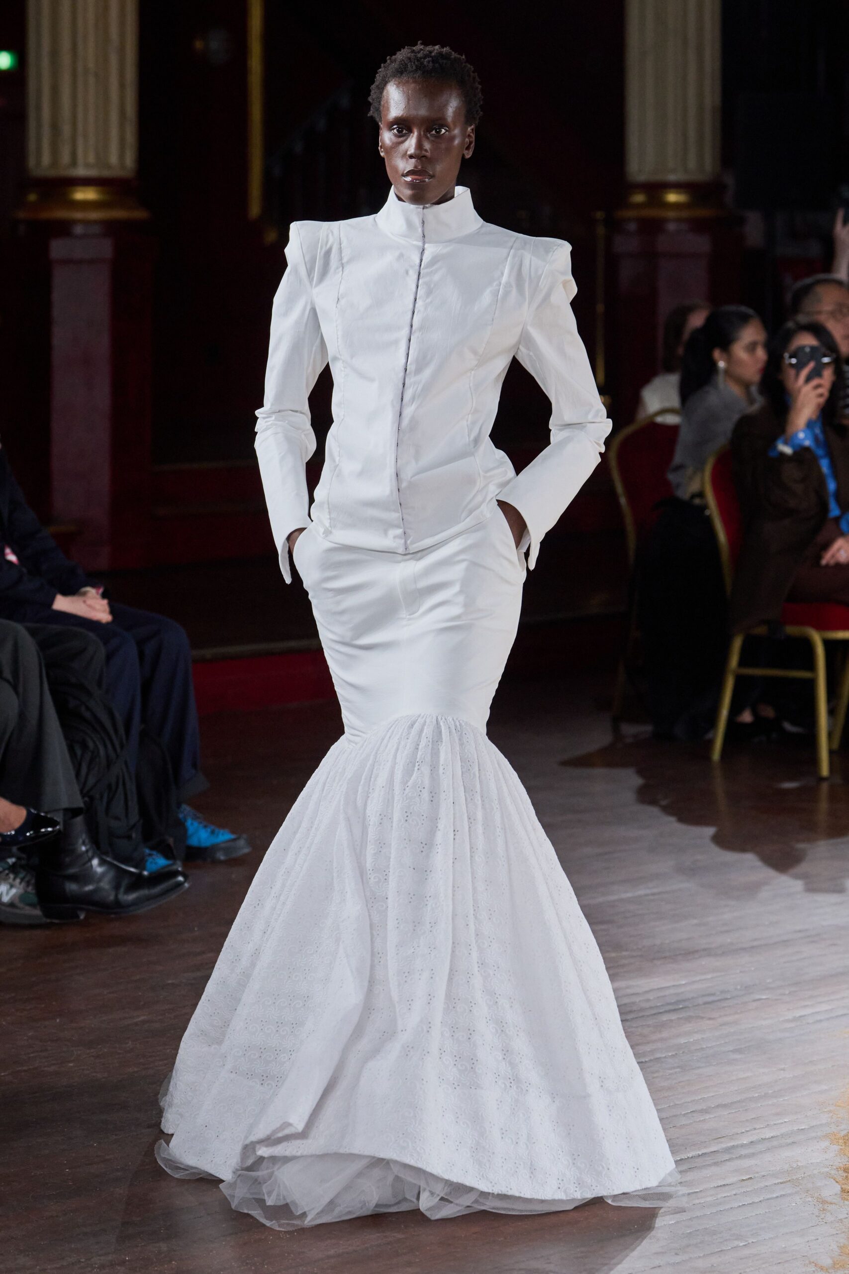 White fitted jacket with a high stand collar and pointed shoulders. Smooth white skirt breaking into an eyelet flounce over tulle. Soft netting visible at the hem. For crossing a ballroom floor after the vows, hands still in pockets.