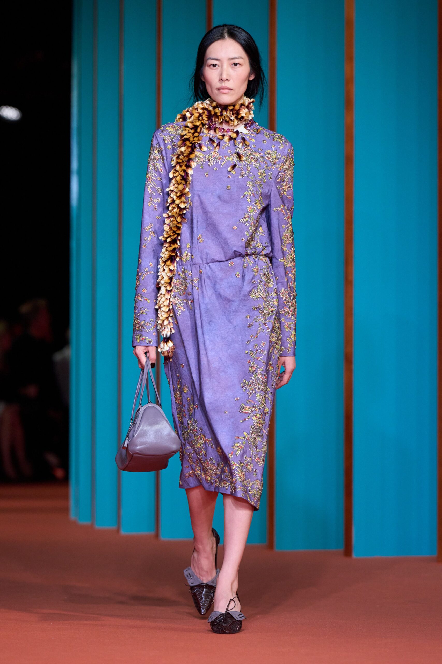 Tiger-shell garland scarf looped tight, one tail dropping past the knee. Lavender long-sleeve dress with scattered metallic floral embroidery and a side tie. Soft lilac leather handbag with rounded gussets. Black woven leather flats with pale bows set off-center. Walking into a hotel atrium still carrying salt from the ferry.
