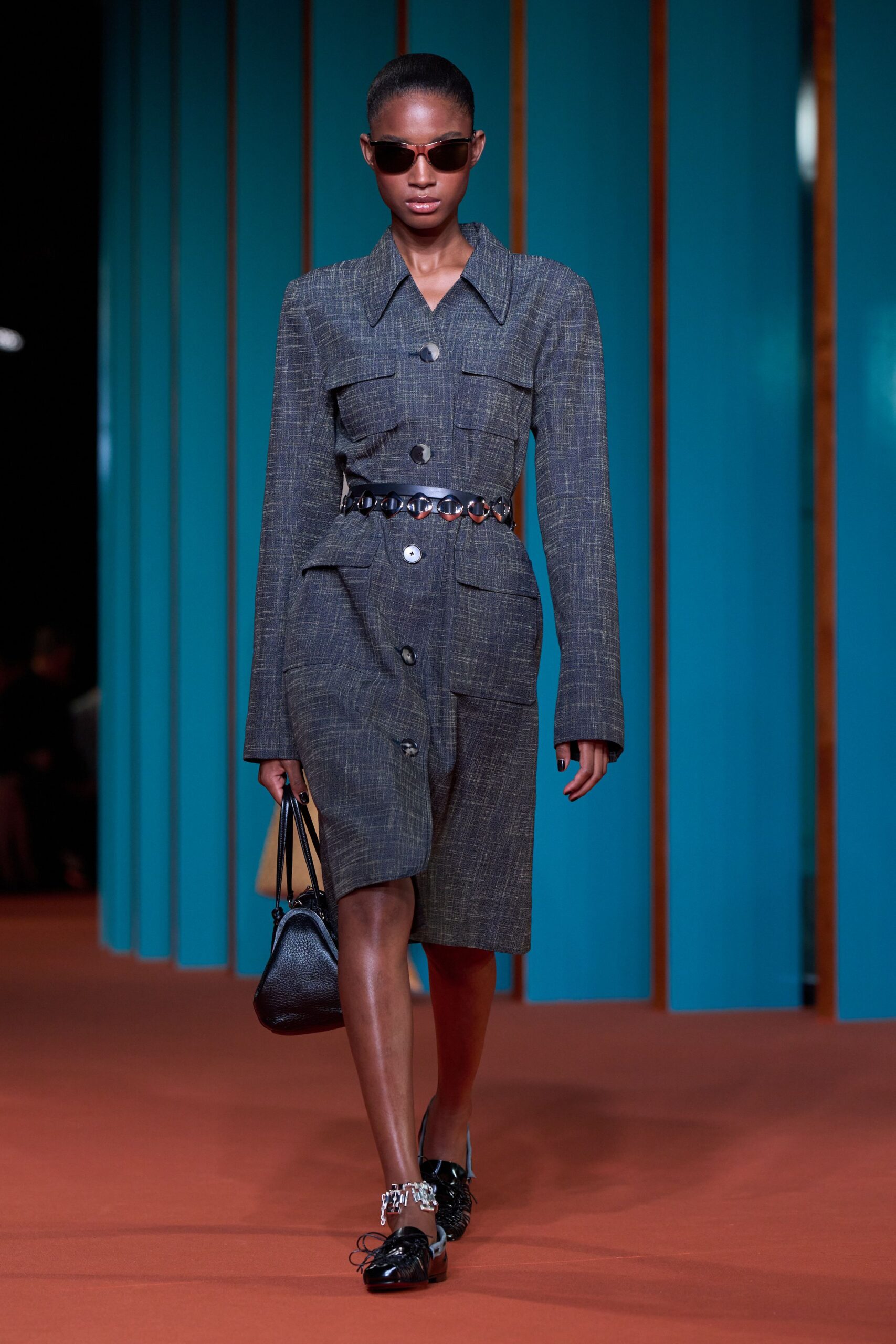 Brown cat-eye sunglasses with narrow temples. Charcoal crosshatch shirt dress, broad collar and oversized horn buttons. Waist belt linked with oval gunmetal discs. Black pebbled leather bucket bag. Black patent lace-up flats with a chunky chain ankle strap. For crossing a civic plaza before the hearing starts.
